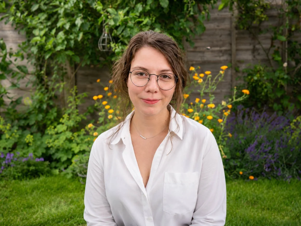 A woman with glasses, wearing a white blouse, sitting outdoors in a garden with green plants, yellow flowers, and purple flowers in the background.