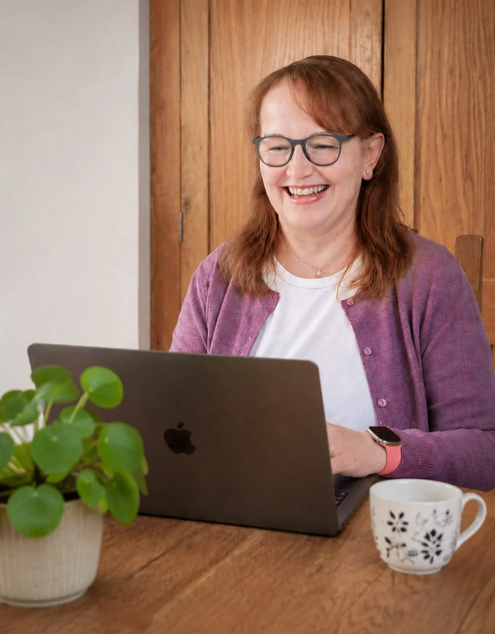 A woman with red hair, glasses, and a white shirt, smiling while using a laptop at a wooden table, with a potted plant and a mug nearby, and wooden cabinets in the background.