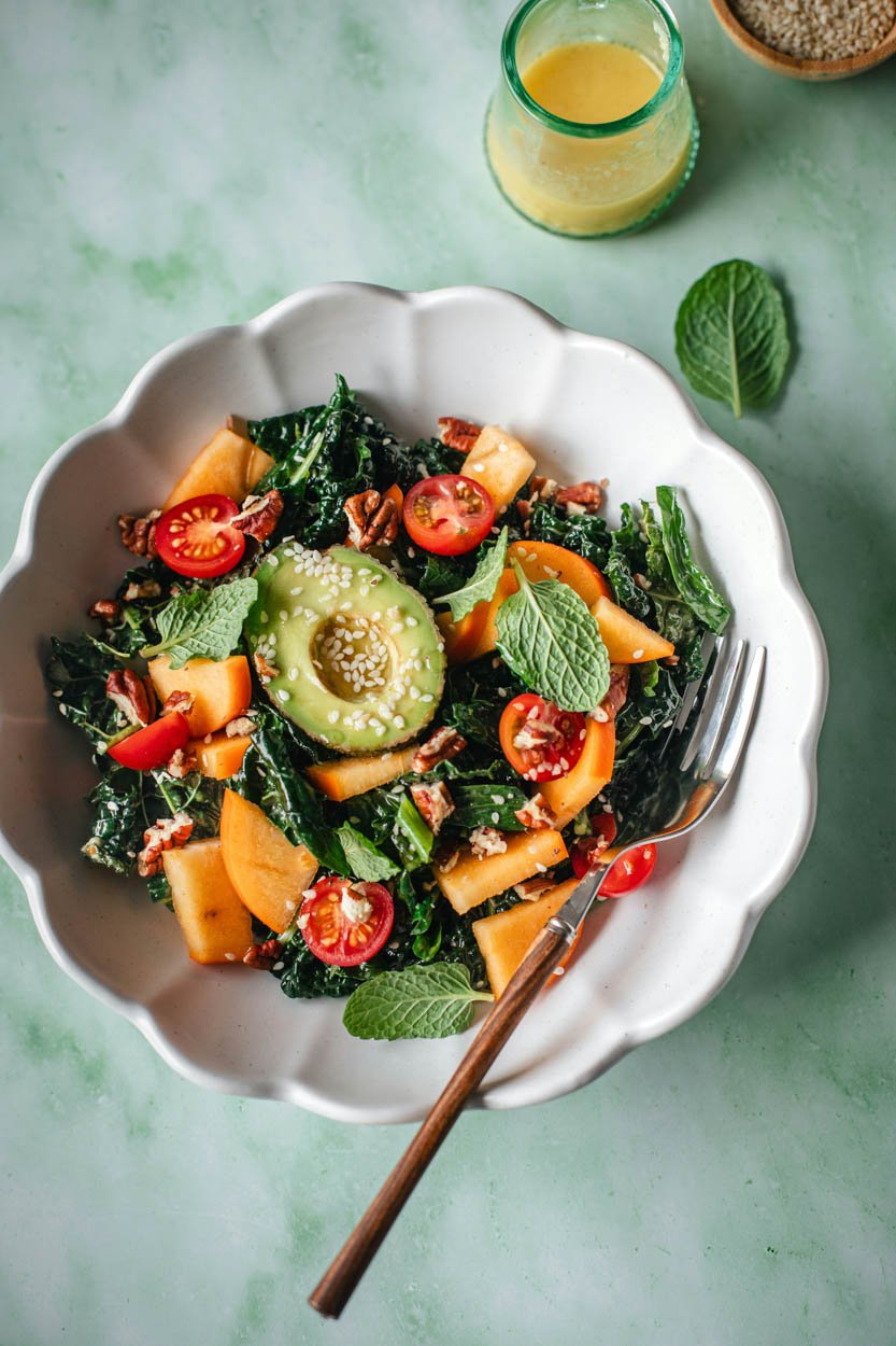 A fresh salad with kale, cherry tomatoes, cantaloupe, avocado slices, walnuts, and fresh mint leaves served in a white scalloped bowl, with a fork on the side, on a green marble surface, accompanied by a glass of lemon or lime beverage and a small bowl of quinoa.
