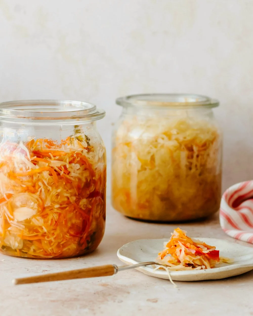 Two glass jars filled with homemade fermented sauerkraut, with a small plate of sauerkraut and a fork in front. A striped cloth is partially visible to the right.