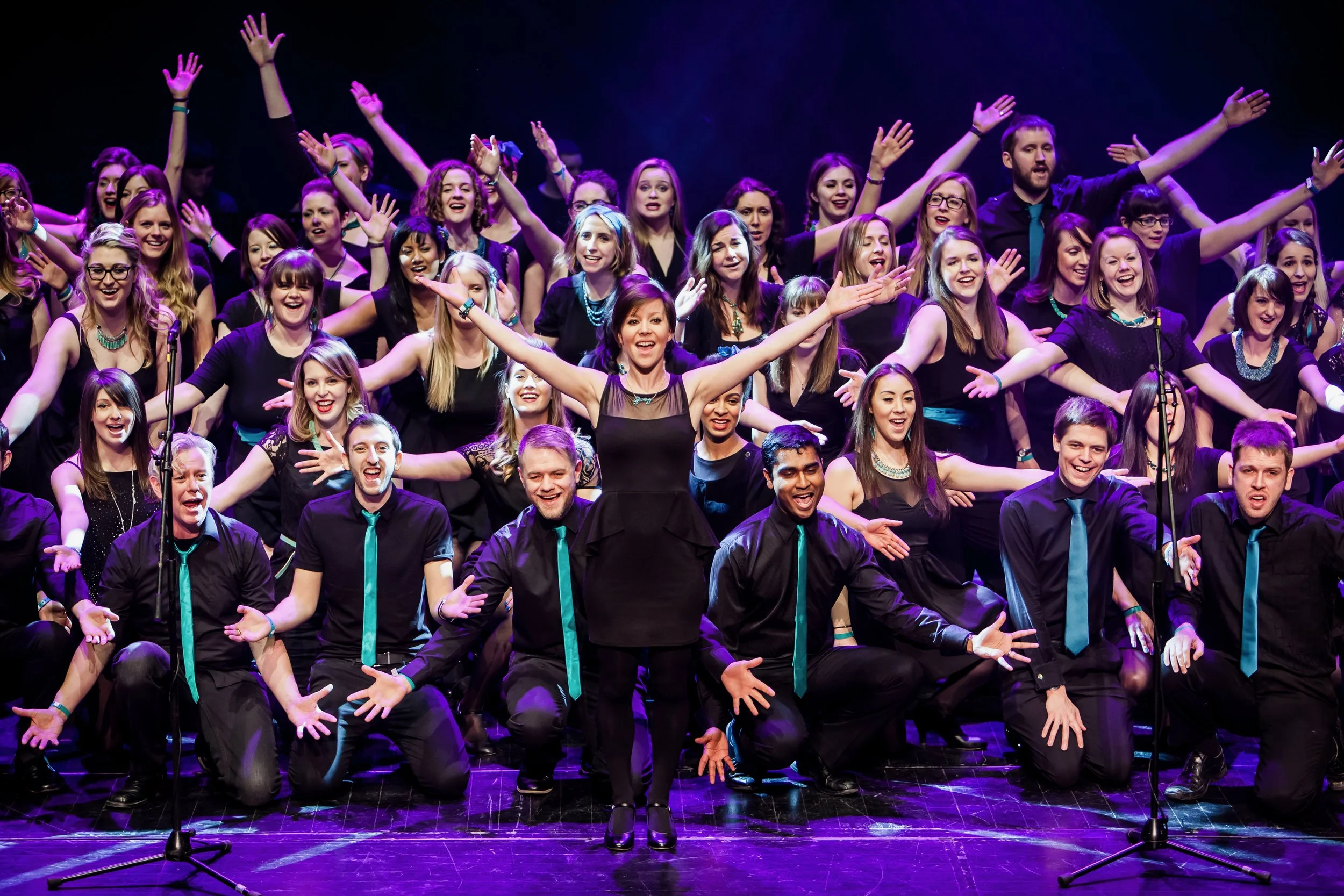 The Starling Arts community choirs pose, hands raised with smiles on their faces, wearing black clothes with teal accessories.