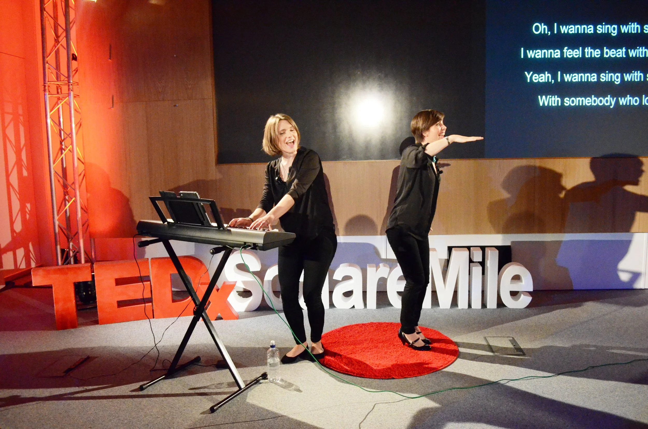 Anna Shields (at the piano) and Emily Garsin stand on stage in front of giant letters spelling TEDx Square Mile