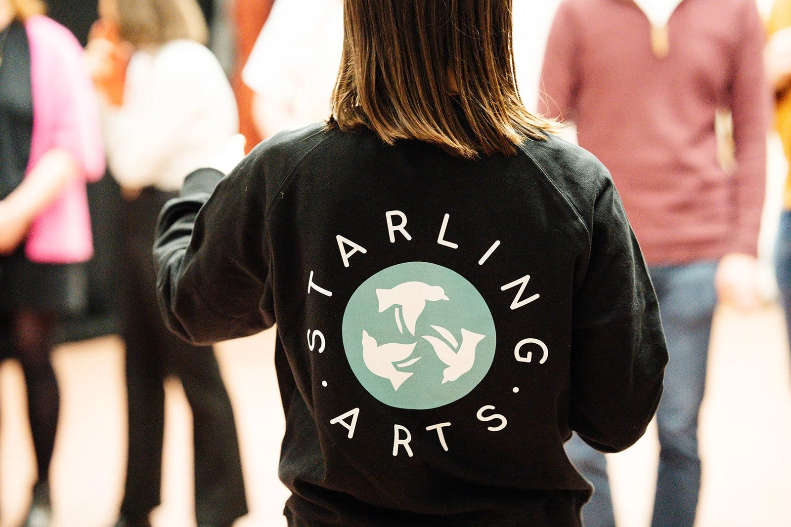 A Starling Arts facilitator wearing a branded black jumper, facing a group during an Applied Musical Theatre community workshop