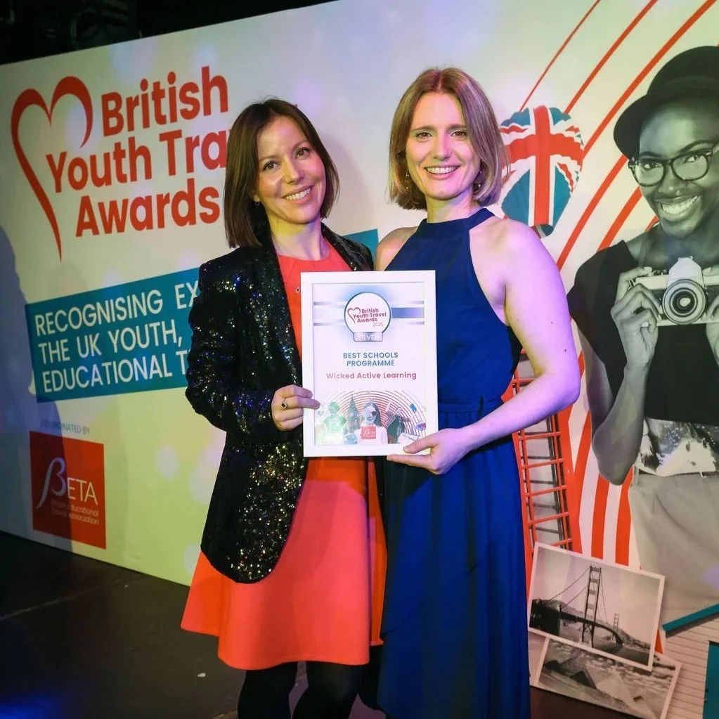 Emily & Anna smiling on stage while holding a framed silver "Best Schools Programme" award from the British Youth Travel Awards 2025 for Wicked Active Learning.