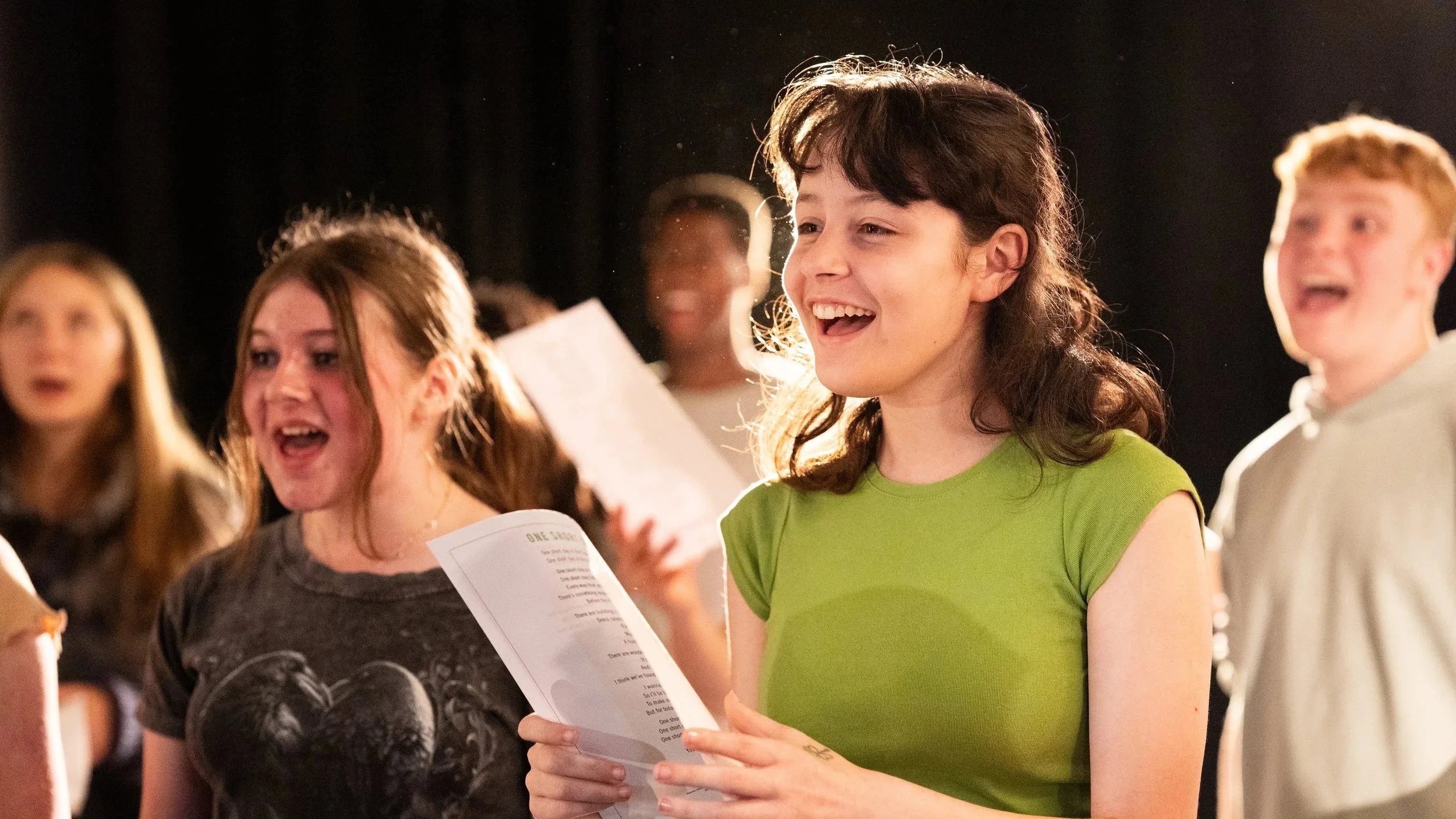 A group of young people singing during a Wicked workshop. In the foreground, a girl in a lime green t-shirt with brown hair