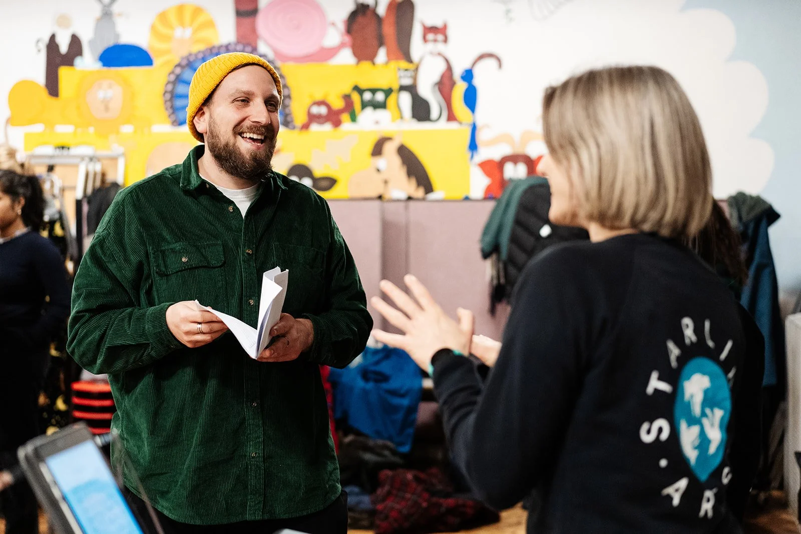 A man in a green shirt and yellow knitted hat shares a joke with a Starling Arts facilitator
