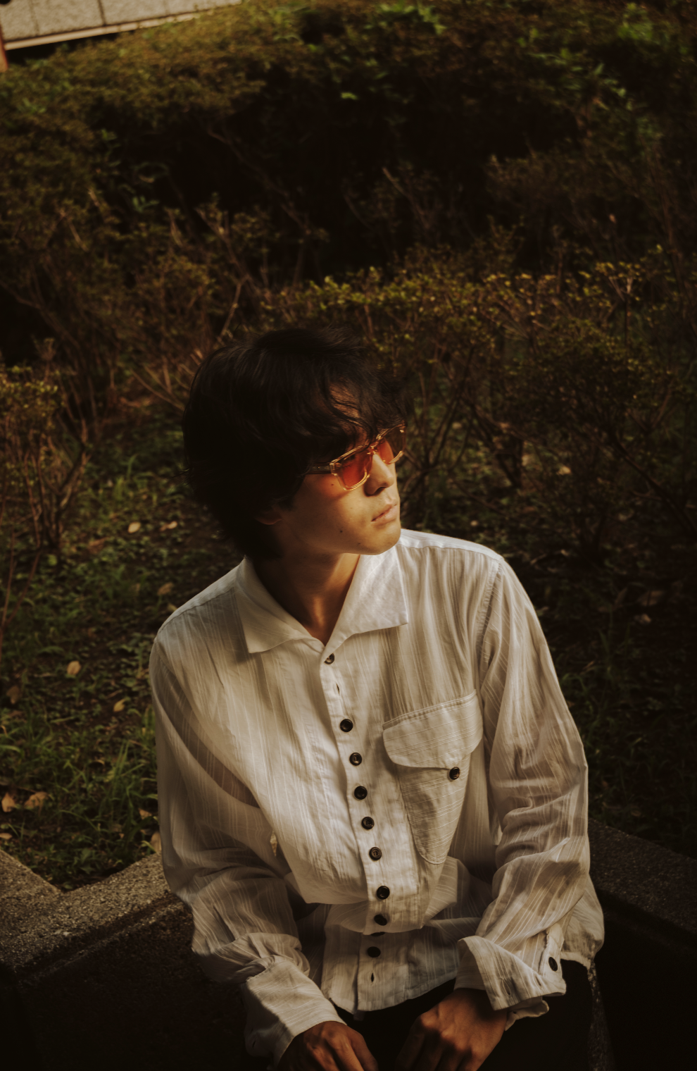 A young man with black hair wearing sunglasses and a cream-colored button-up shirt sitting on a concrete bench outdoors during the evening.