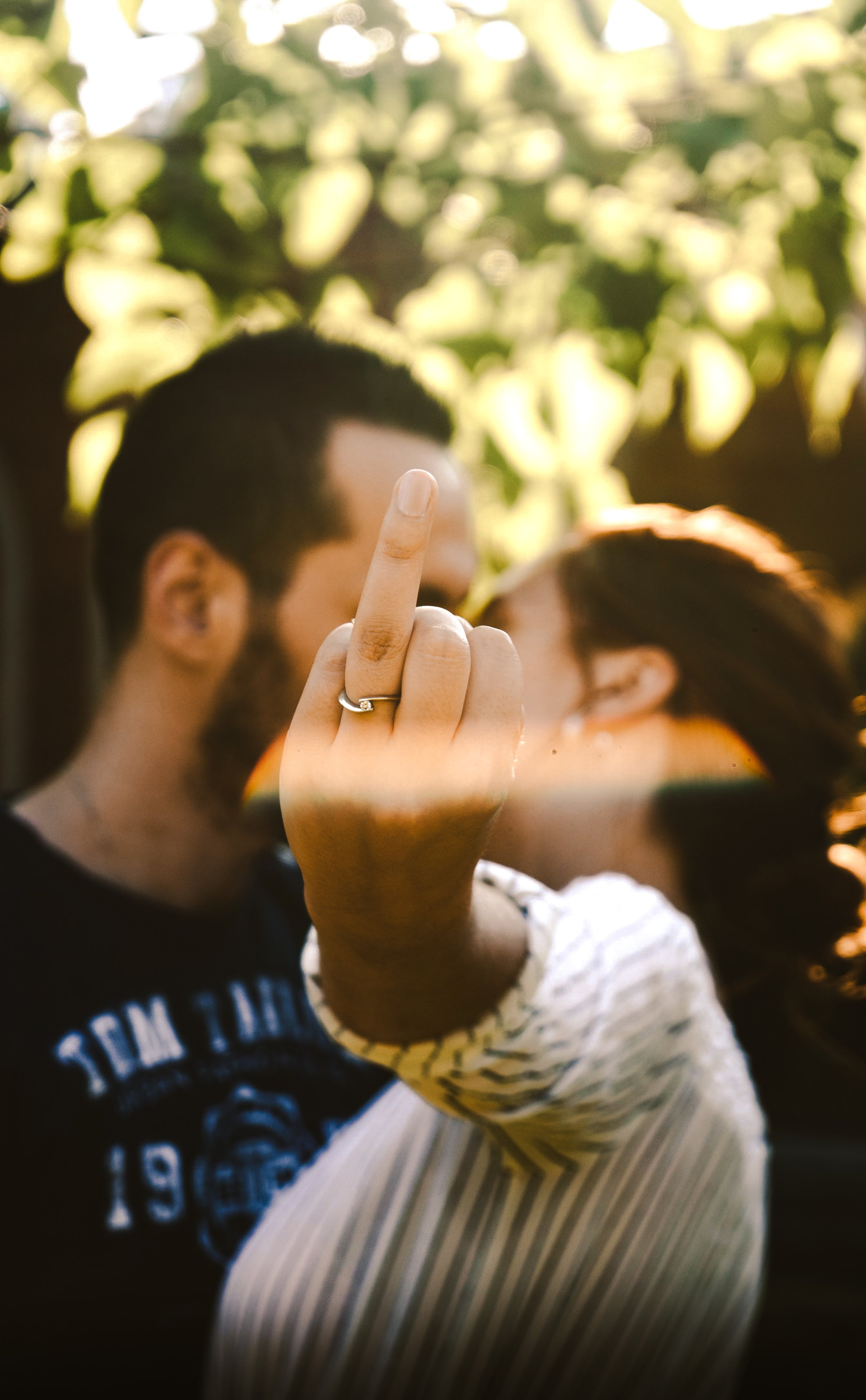 Person displaying the ring finger, wearing a ring, in front of a blurred couple at a gathering outdoors with yellow sunlight.