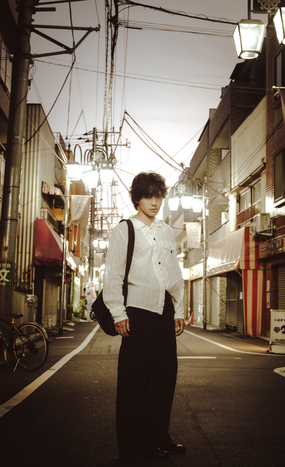 A young man stands in the middle of a quiet urban street at dusk, wearing a white button-up shirt and black pants, carrying a black shoulder bag, with city buildings and streetlights in the background.