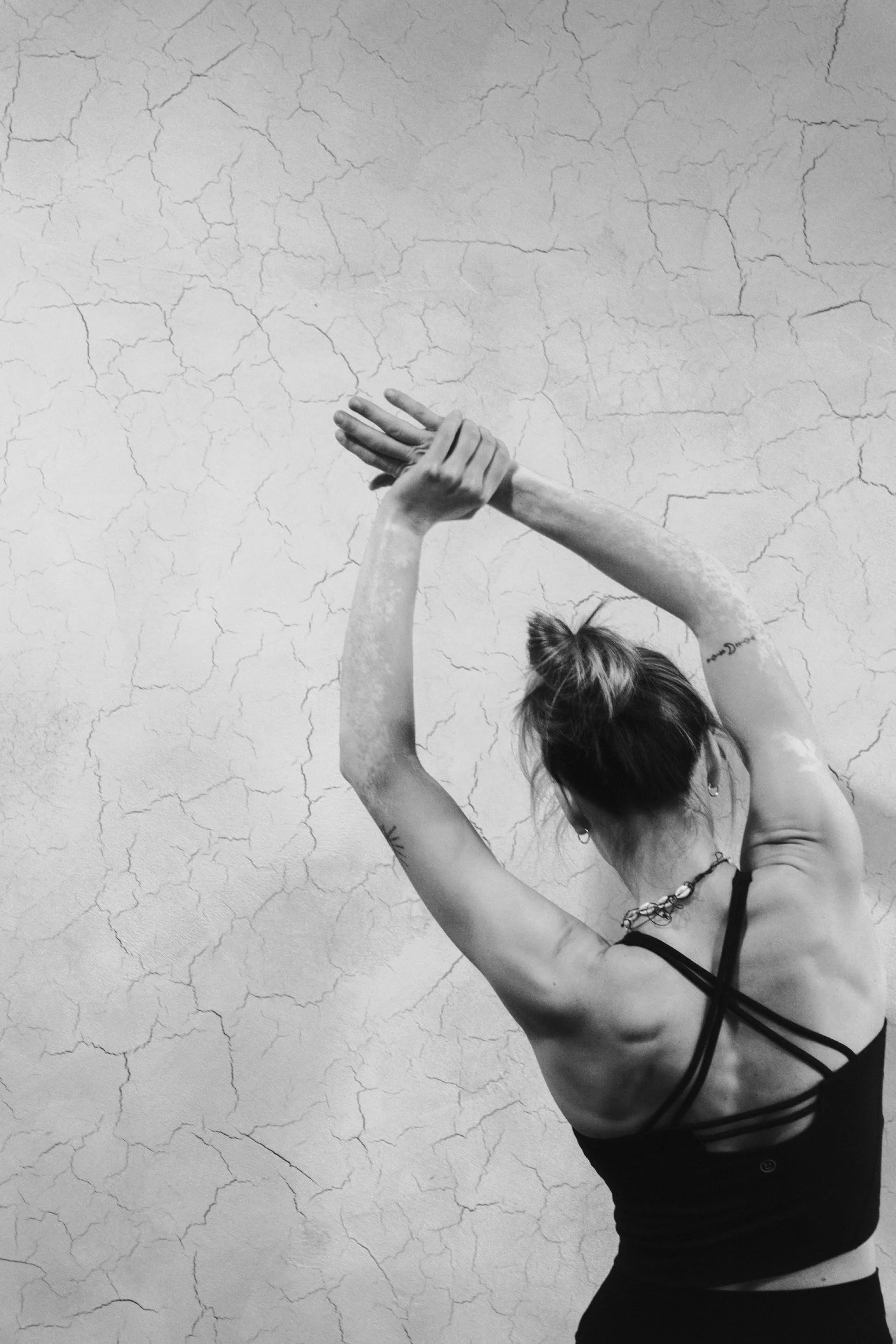 Black and white photo of a woman practicing yoga or stretching, with her arms raised and hands pressed together, against a cracked wall background, wearing a sports bra and leggings, with her hair in a bun.