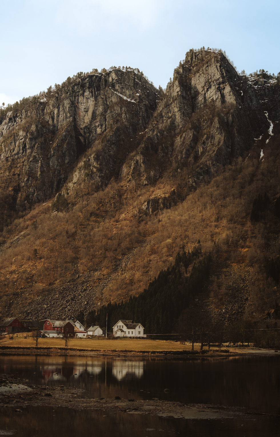 View of mountains with rocky cliffs and sparse trees, a small cluster of houses near a reflective lake.