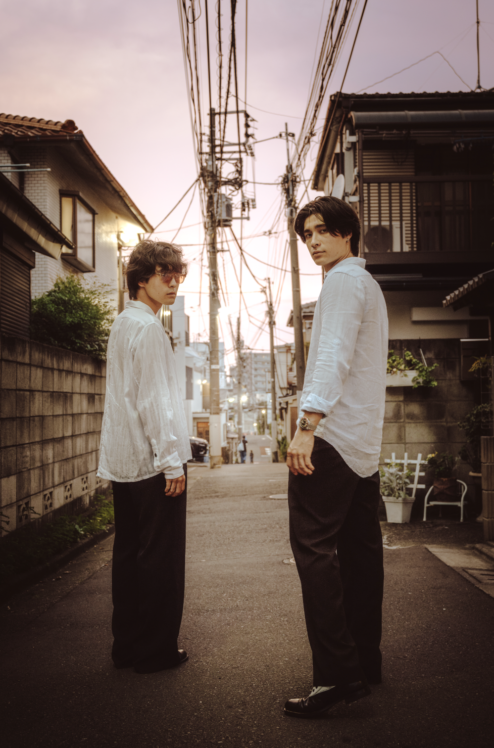 Two young men standing in an alley at sunset, wearing casual white shirts and dark pants, with utility poles and residential buildings in the background.