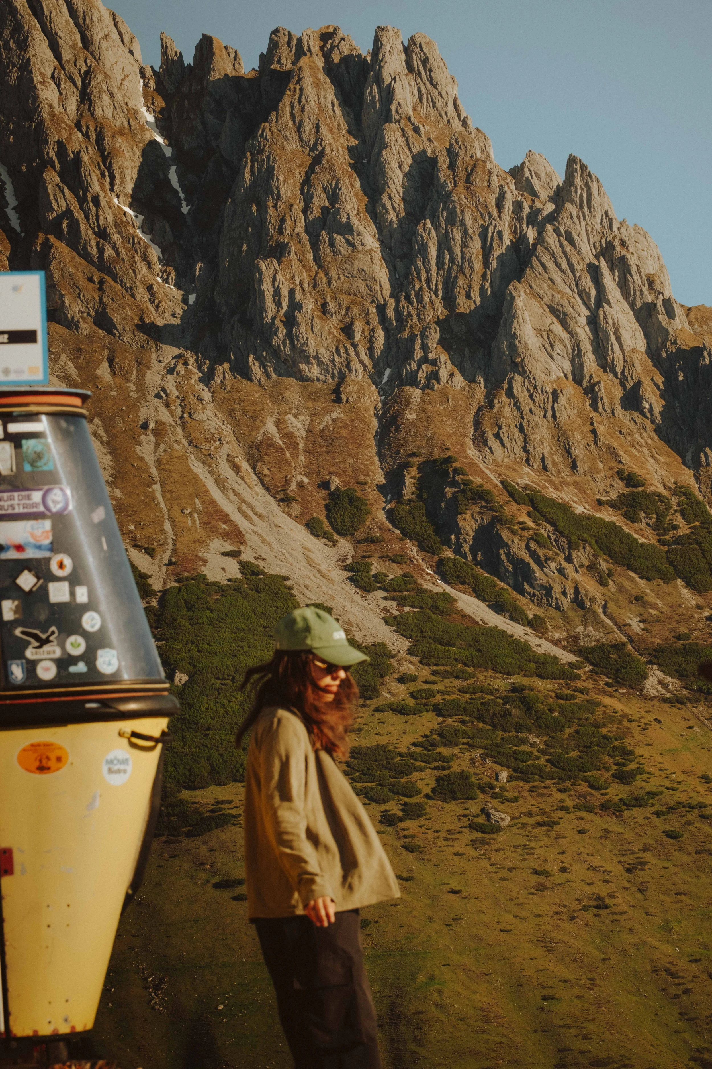 A woman walking outdoors near a mountain with rocky peaks and sparse vegetation, wearing a green cap and sunglasses.