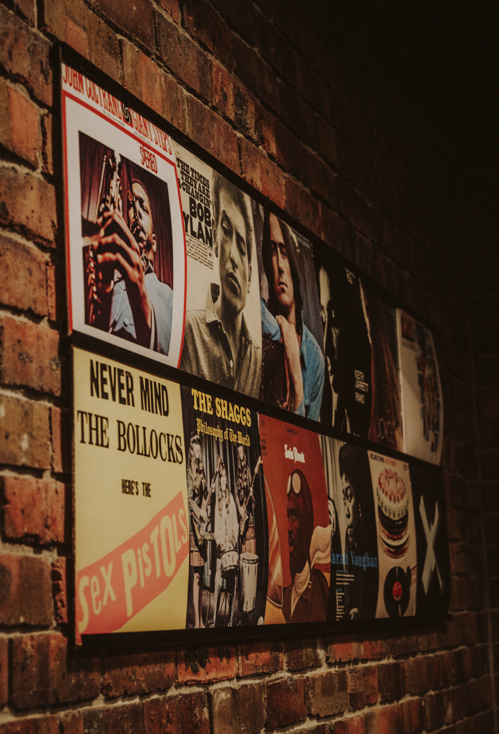 A collage of vintage vinyl record album covers mounted on a brick wall, featuring notable artists such as Bob Dylan, The Sex Pistols, The Shaggs, Sarah Vaughan, and The Velvet Underground.