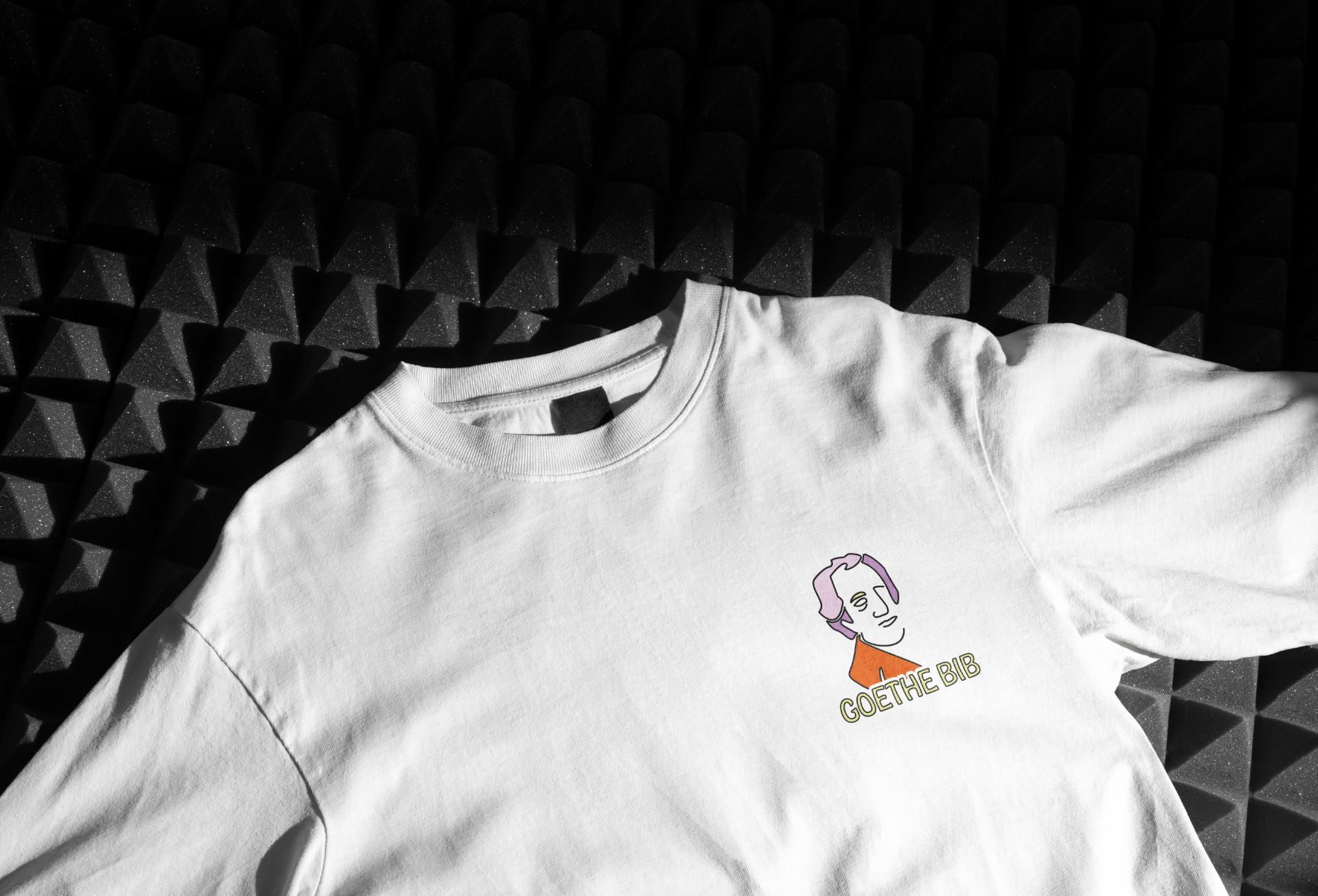 White T-shirt with a colorful graphic of Goethe and the text "GOETHE BIB" on it, lying on a black textured surface.