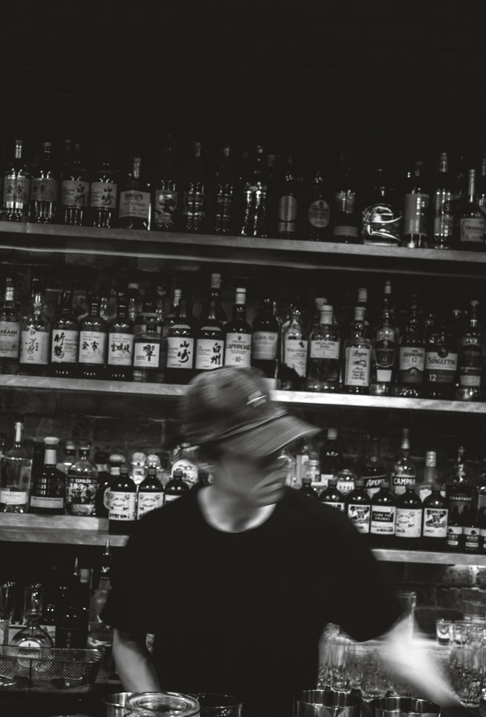A person behind a bar counter with shelves of liquor bottles in the background, wearing a baseball cap and black shirt, in a black and white photo.