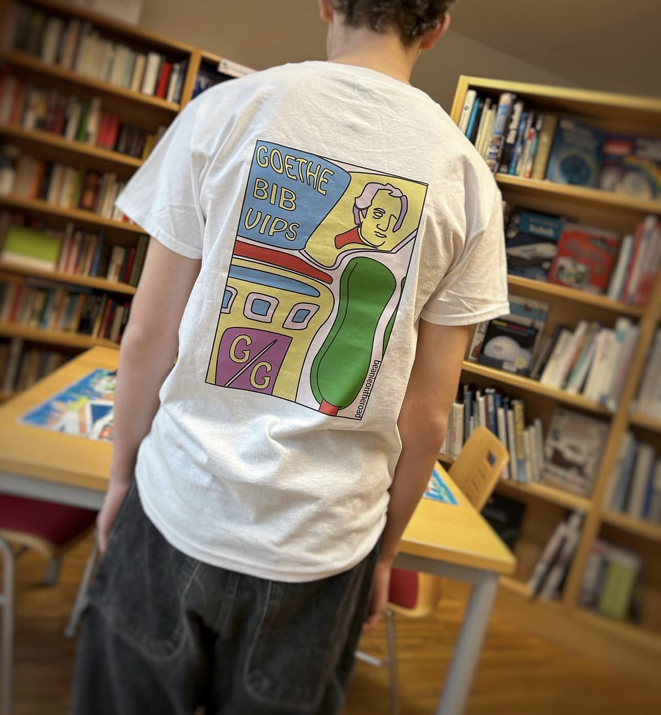 Person wearing a white T-shirt with a colorful, abstract graphic design and text, standing inside a bookstore with wooden bookshelves filled with books.