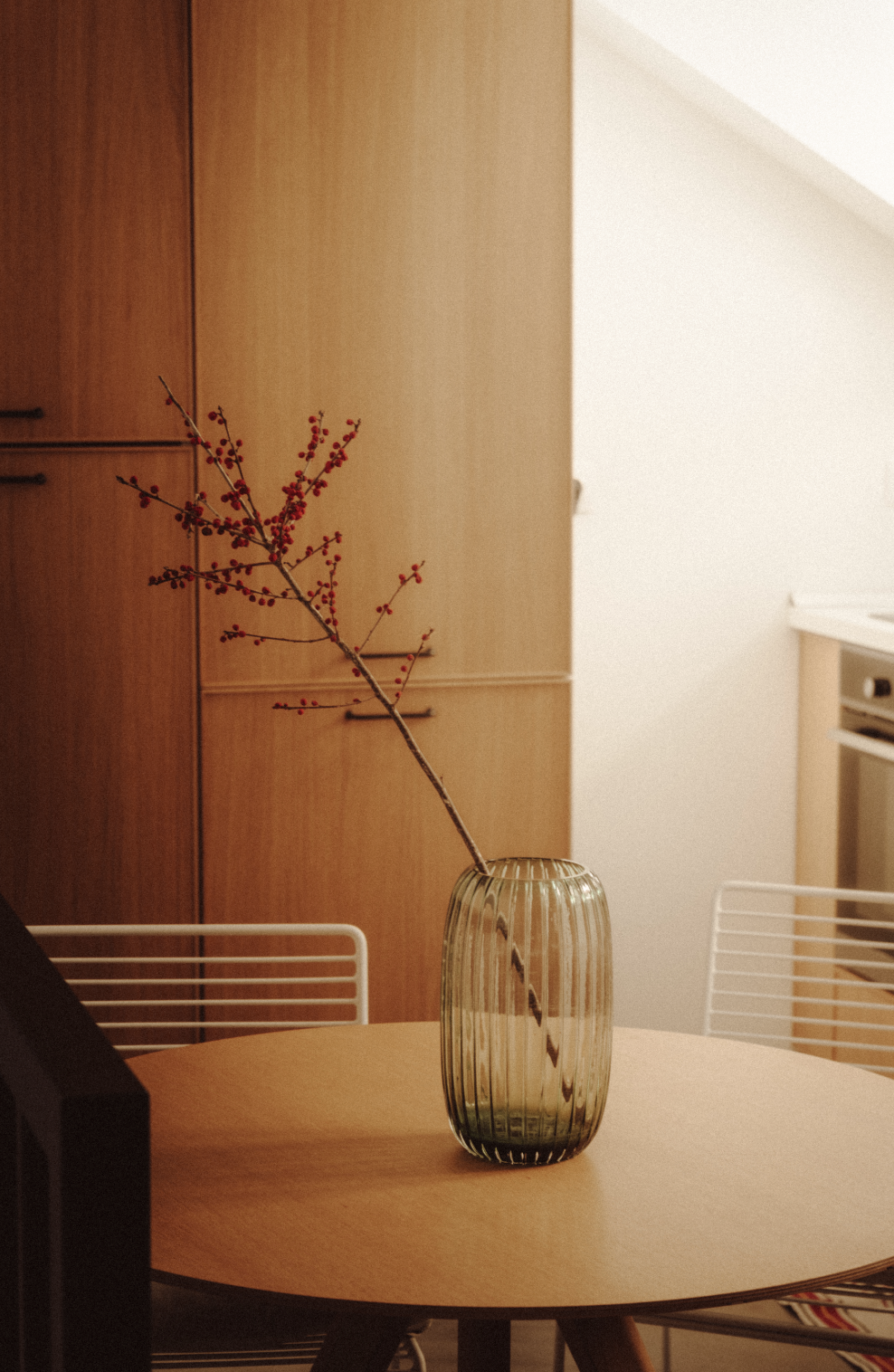A small, striped glass vase with a long, thin branch of red berries, placed on a round wooden table in a cozy kitchen or dining area.