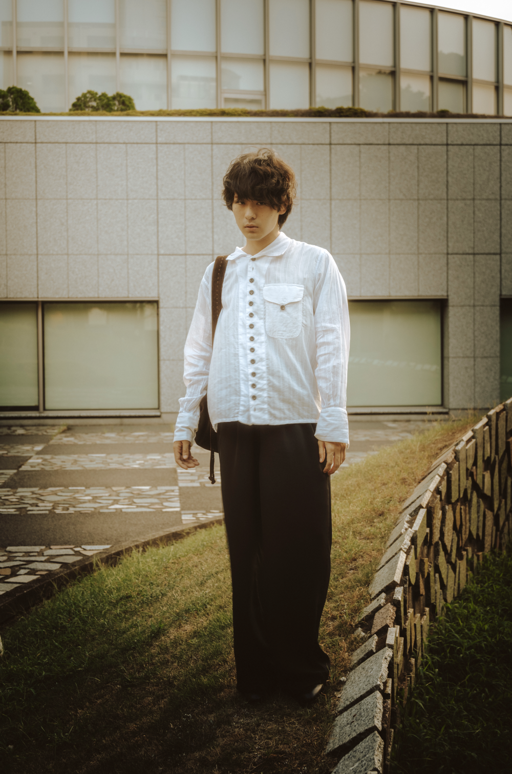 Young man standing outdoors on a grassy area with a modern building in the background, wearing a white button-up shirt and black pants, carrying a backpack.