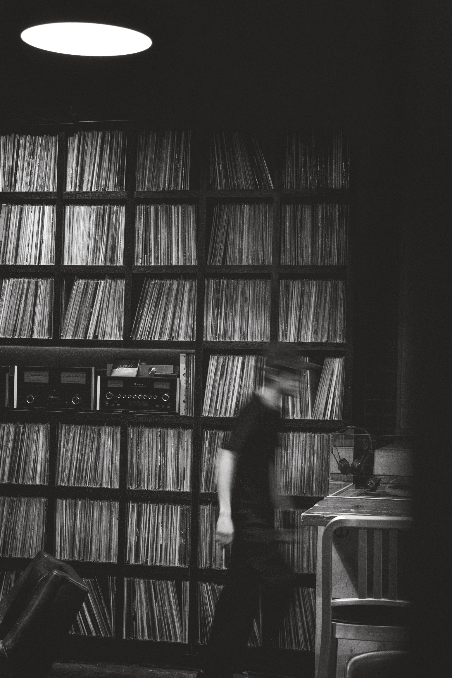 A black and white photo of a room with shelves filled with vinyl records. A person appears to be moving, causing a motion blur, and there's a desk with a chair and audio equipment in the room.