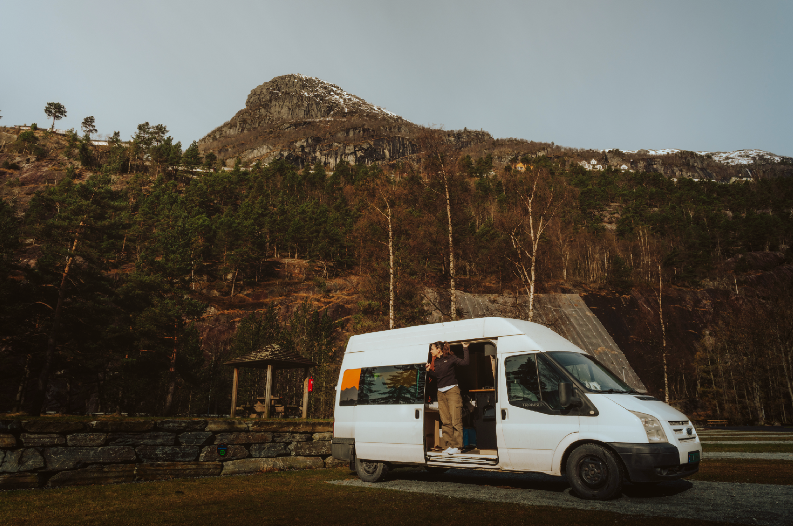 A person standing at the open sliding door of a white van, parked in front of a mountainous landscape with trees and a rocky hillside.
