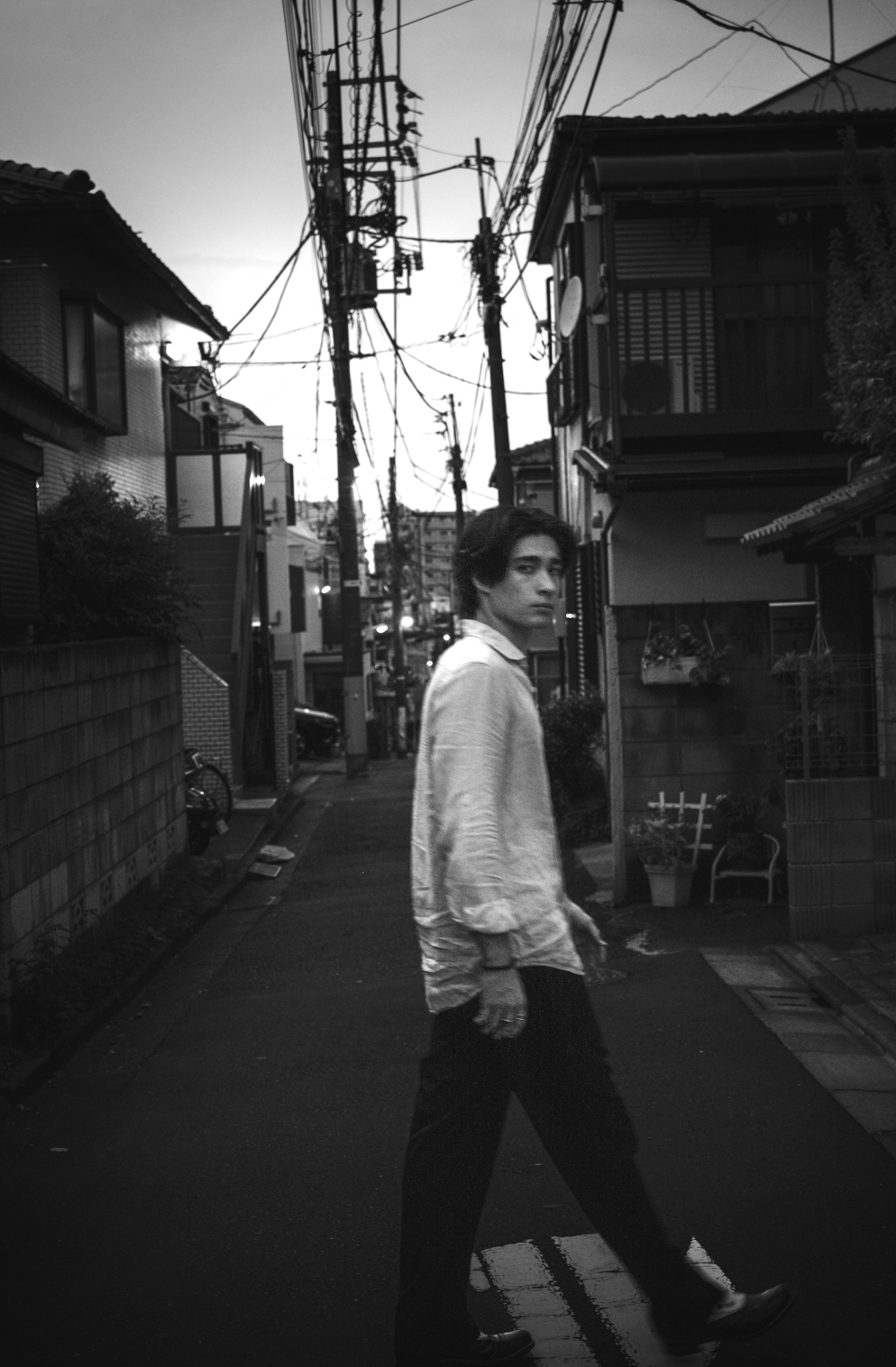 A black and white photo of a young man with dark hair and light-colored shirt crossing a street in an urban neighborhood, with houses, utility poles, and overhead wires visible in the background.