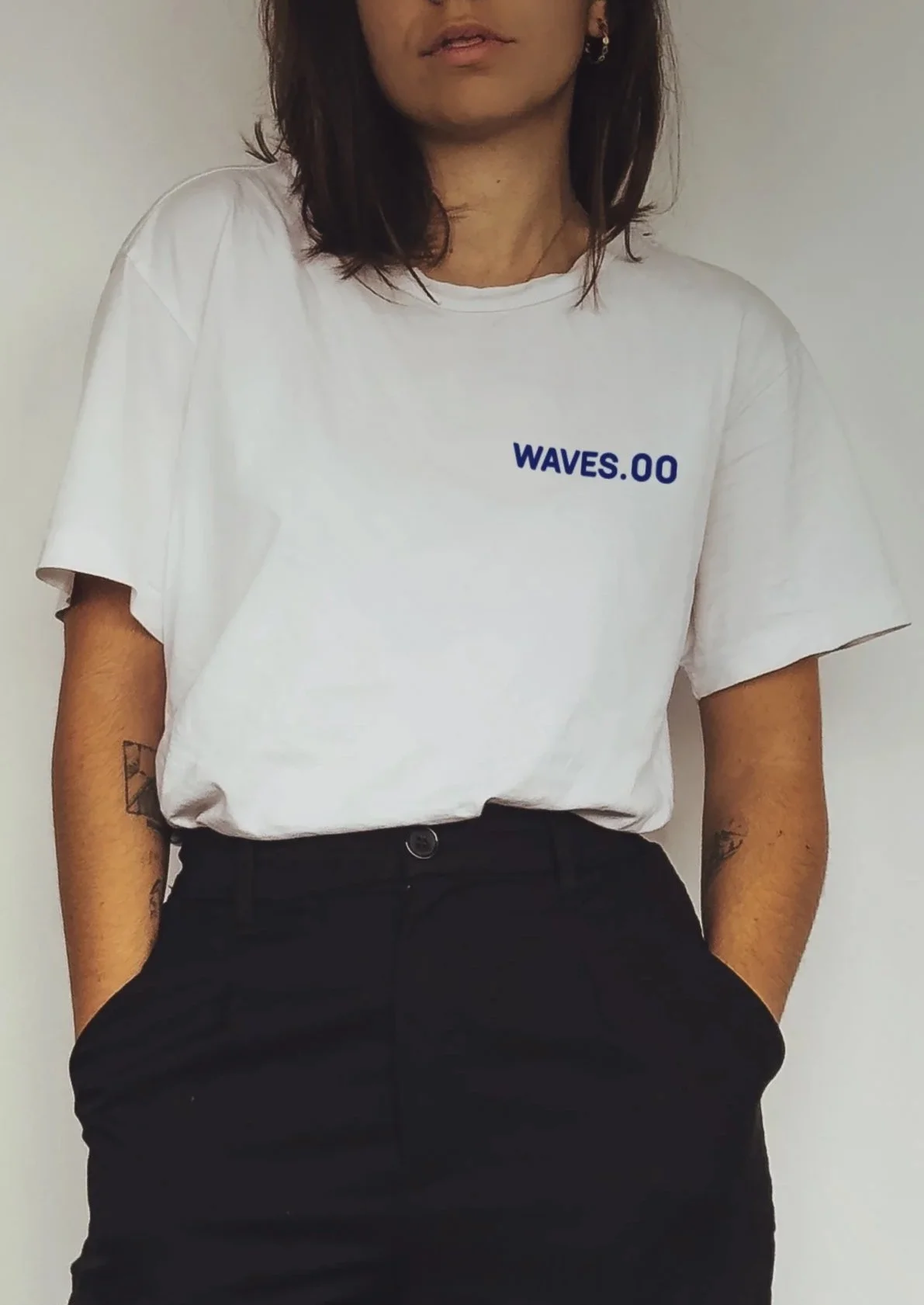 Person wearing a white t-shirt with blue text reading 'WAVES.00', black pants, standing with hands in pockets, with tattoos on arms, against a plain white background.