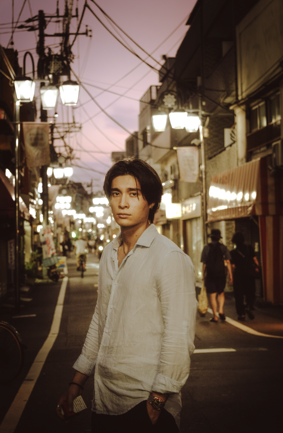 A young man with dark hair and a light-colored shirt standing on a street at dusk with a pinkish sky. The street is lit with street lamps and lined with buildings, signs, and pedestrians.