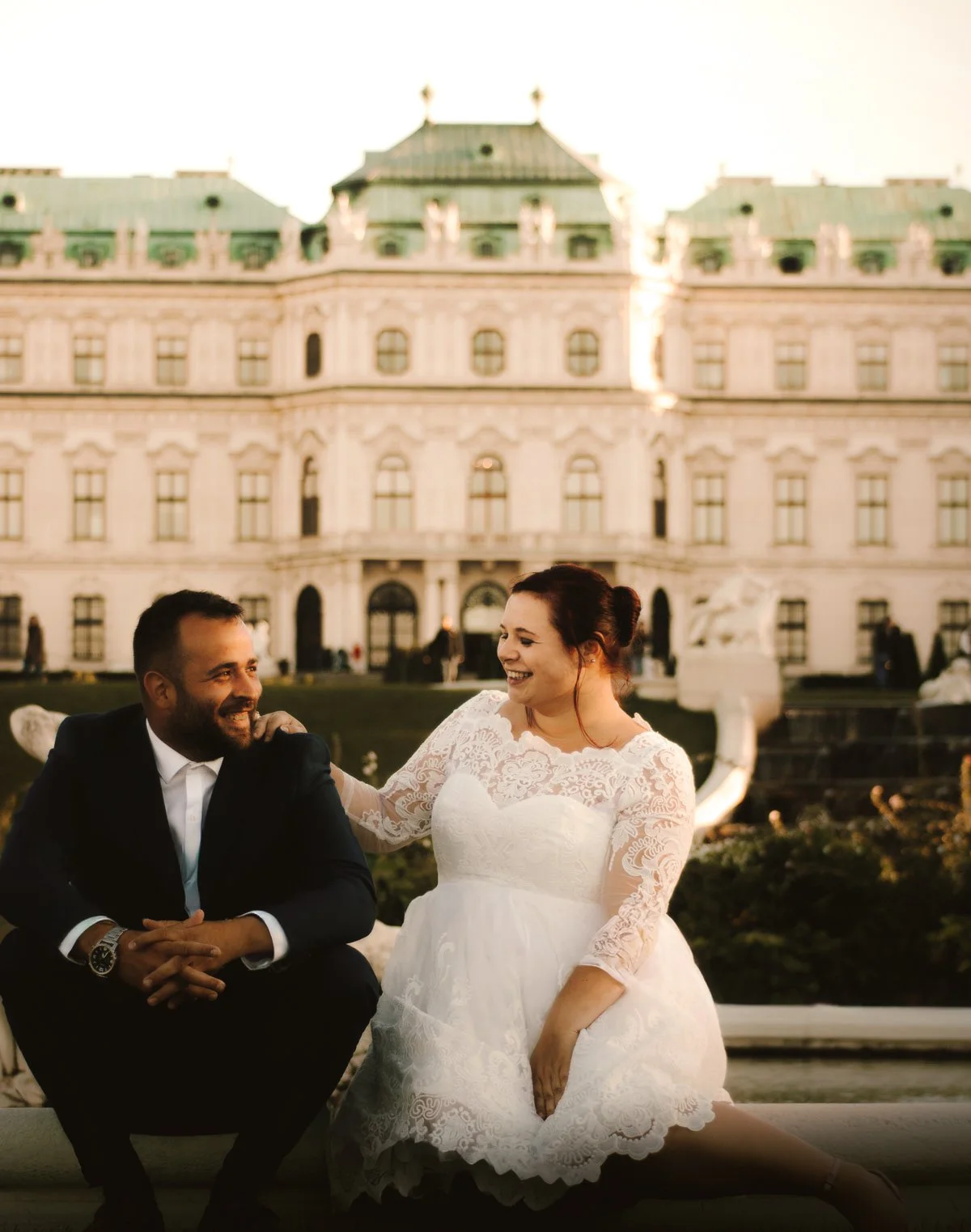 A bride and groom sitting outdoors in front of a large historic building, sharing a joyful moment during their wedding photos.
