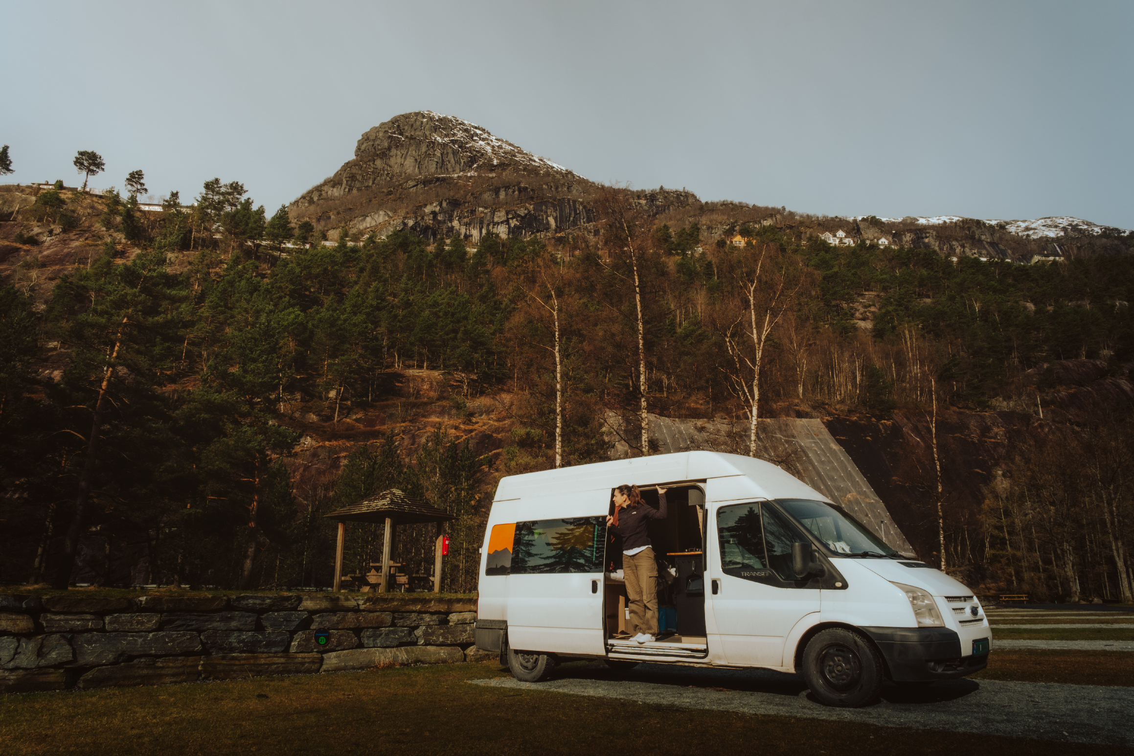 A white van parked outdoors with a woman standing at the open side door, with a mountain with a snow-capped peak and trees in the background.