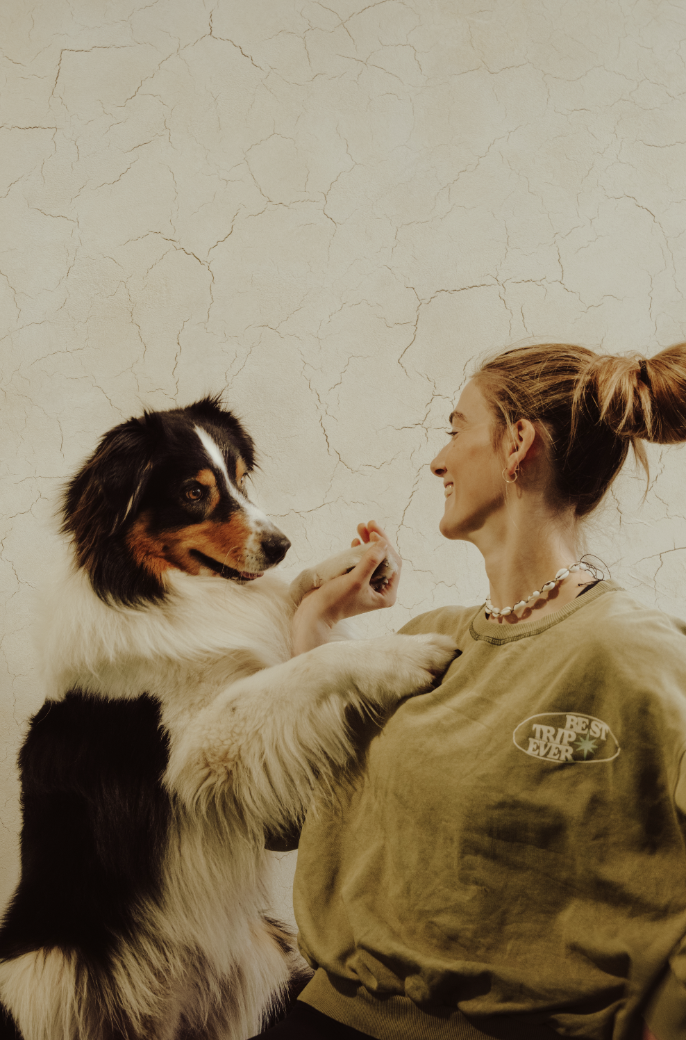A woman with blonde hair in a ponytail smiling and interacting with a black, white, and brown Border Collie dog against a cracked beige wall.