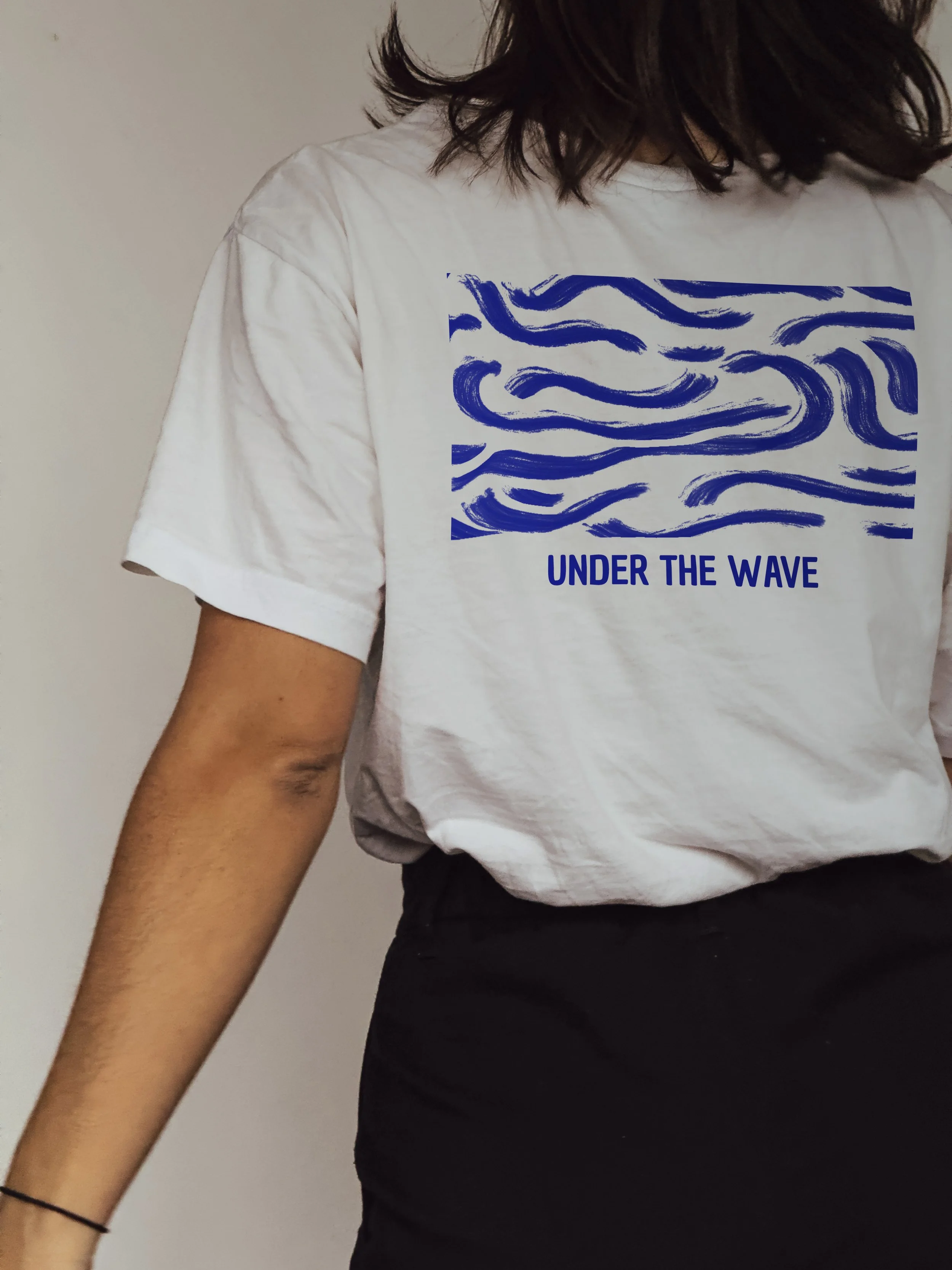 A person wearing a white T-shirt with a blue graphic design of wavy lines and the text 'UNDER THE WAVE' underneath, black pants, and a black bracelet on their wrist.
