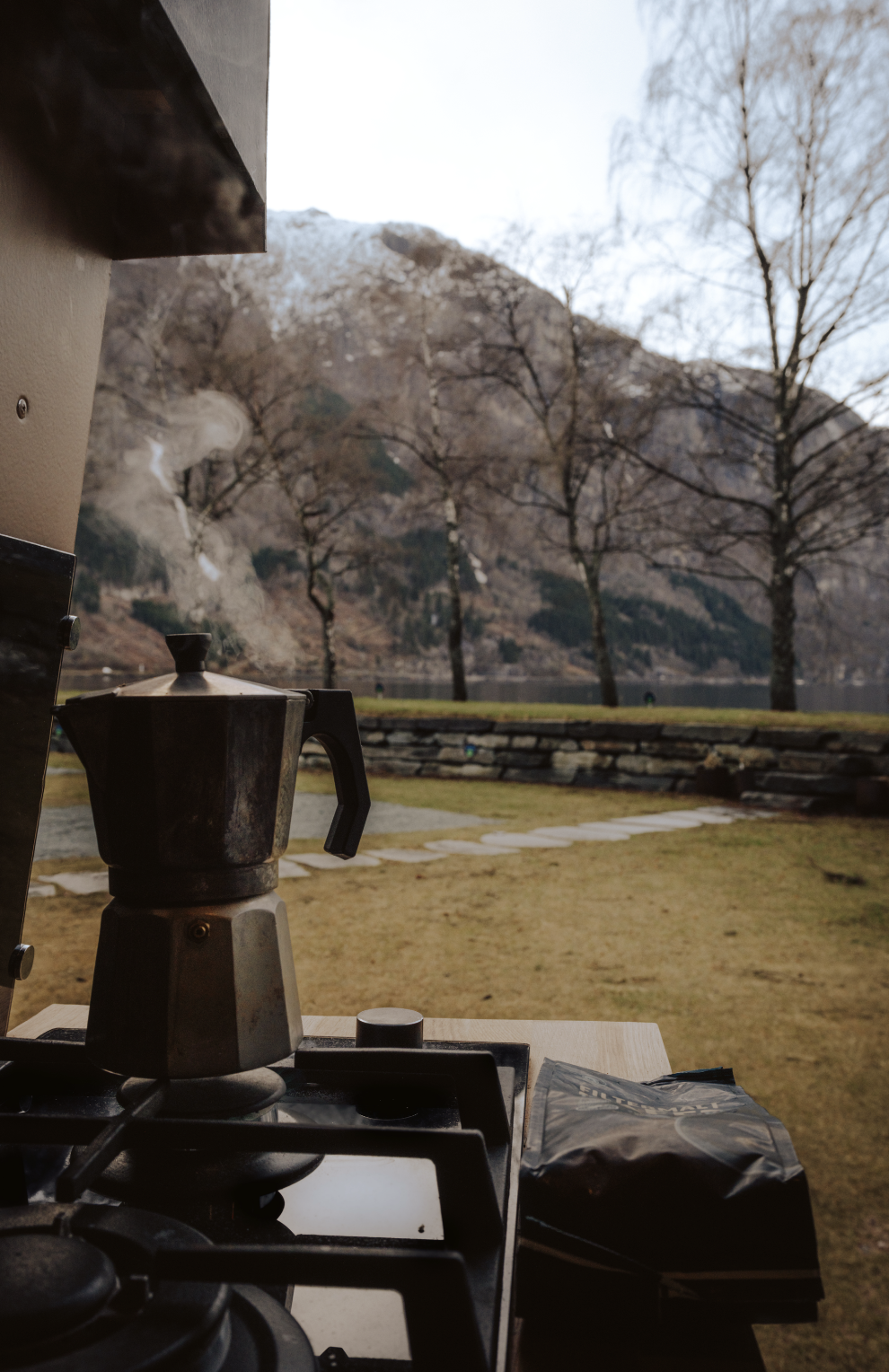 Coffee maker on a countertop outdoors with mountains, leafless trees, and a stone pathway in the background.