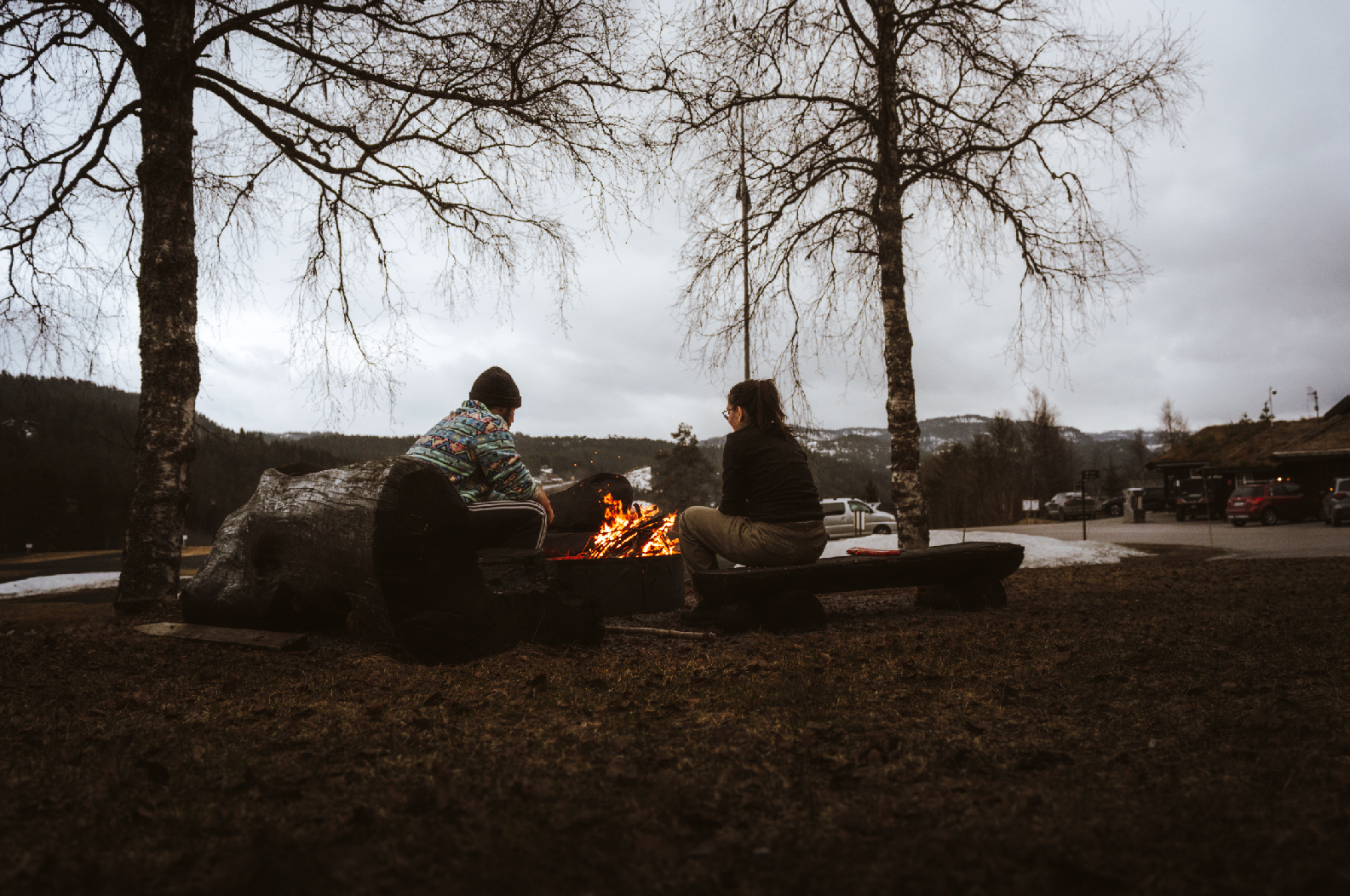 Two people sit by a campfire under leafless trees on a cloudy day in a rural area, with parked cars and mountains in the background.