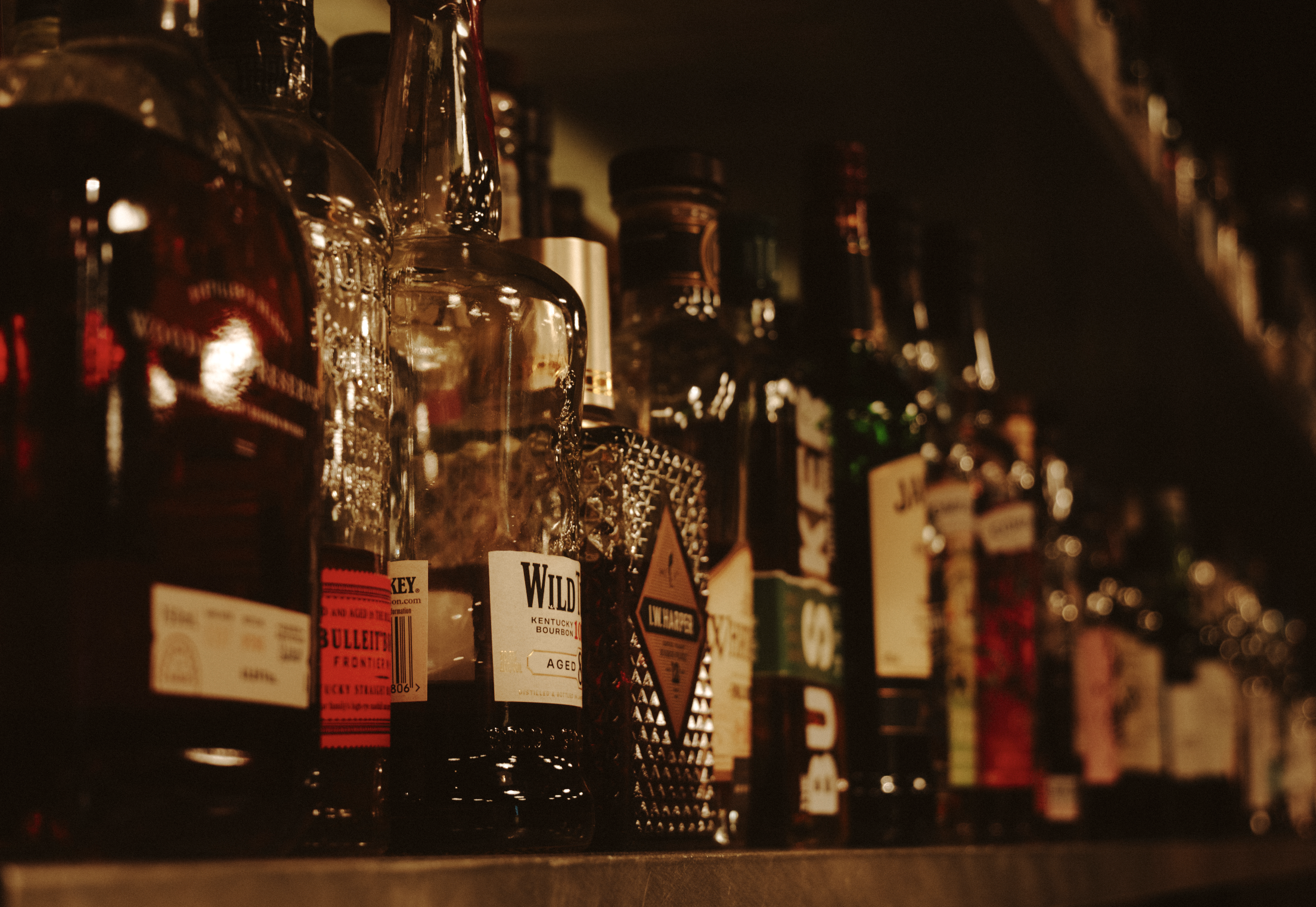 A shelf stocked with various bottles of alcohol, including whiskey and bourbon, in a bar or liquor store setting. The bottles have different shapes, labels, and colors, some with warning and age restriction tags.