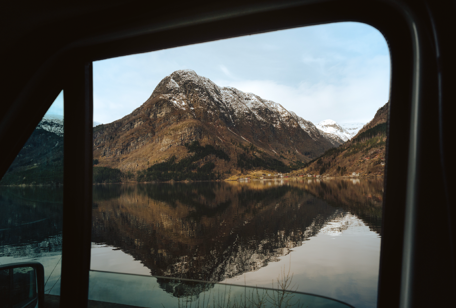 Mountain landscape viewed through a vehicle window, with snow-capped peaks and a calm lake reflecting the mountains.