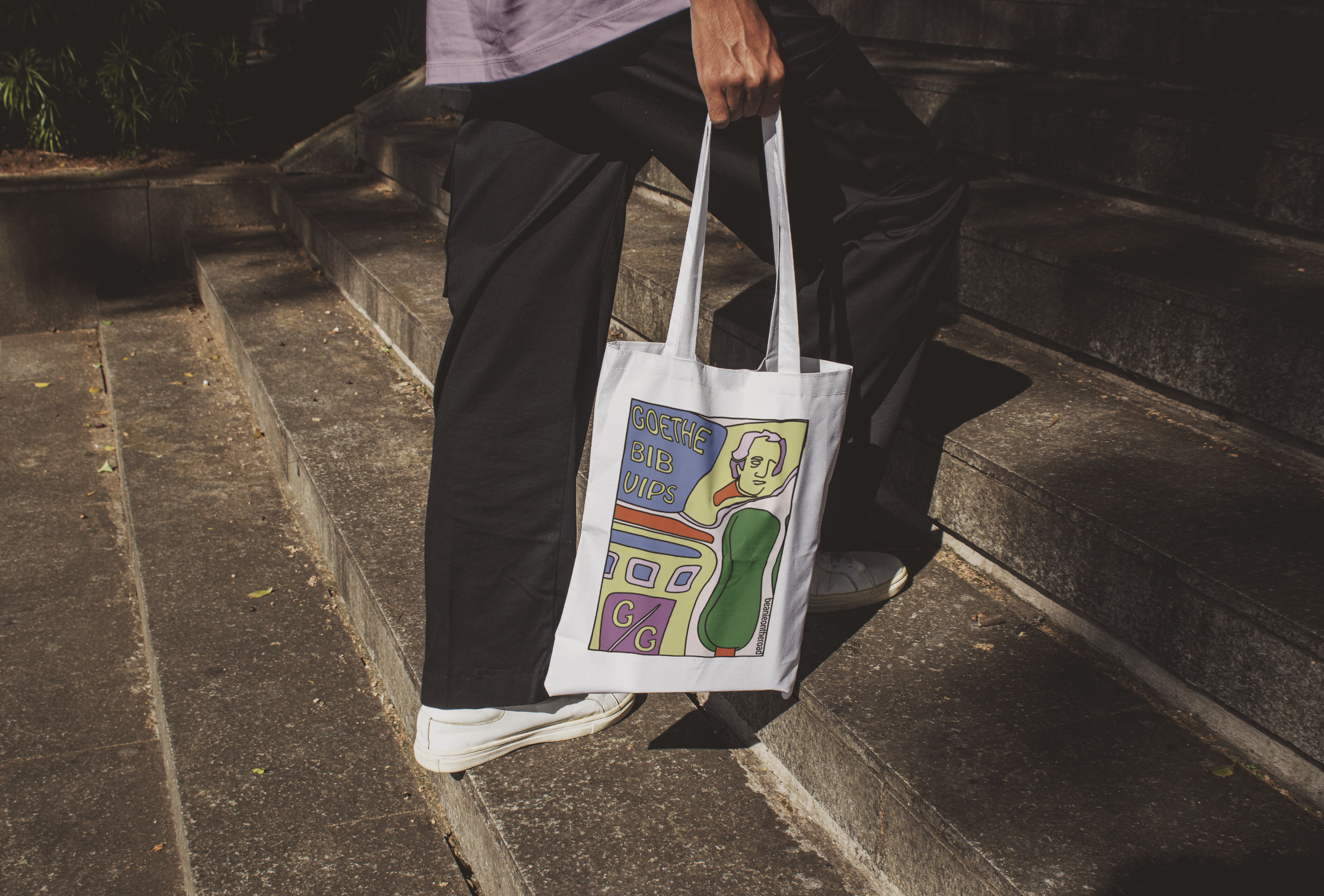 Person sitting on outdoor concrete steps holding a tote bag with a colorful design, wearing white sneakers, black pants, and a purple shirt, with greenery and shadows in the background.