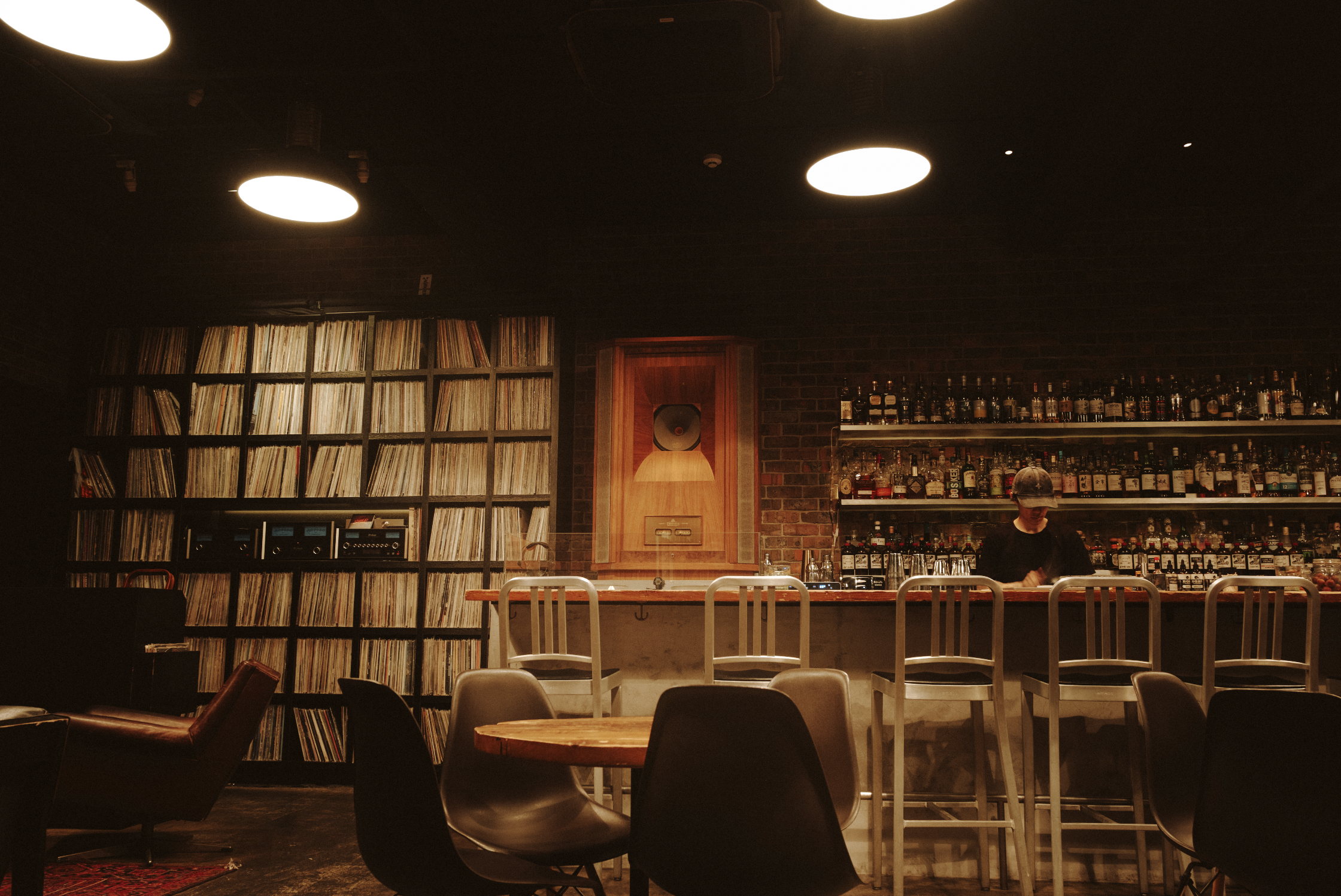 A dimly lit bar with a bookshelf of vinyl records on the left, a bar counter with a bartender and liquor bottles on the right, and seating in the foreground.