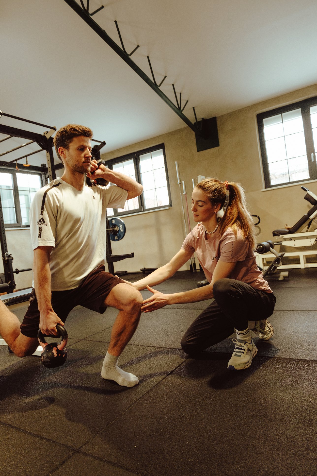 A man is holding a kettlebell in one hand and a phone in his other hand while doing a lunge exercise, guided by a woman in a gym. The woman is kneeling and supportive as he performs the exercise. The gym has windows and various workout equipment.