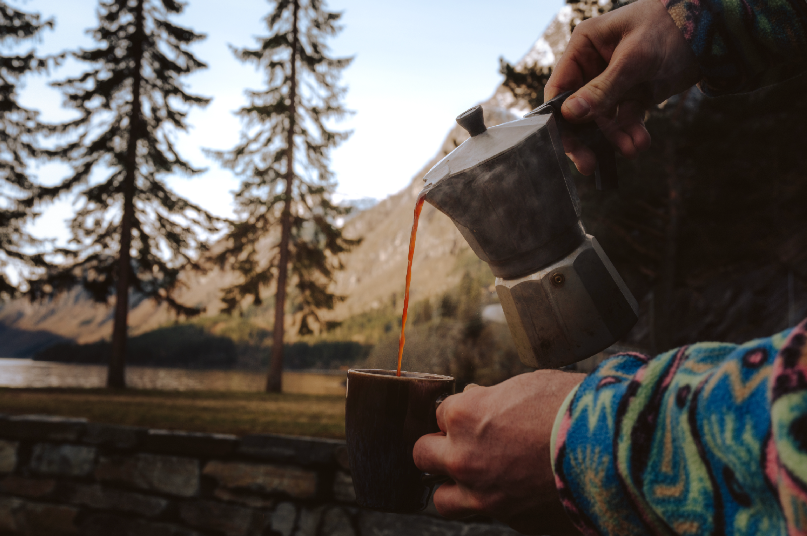 Person pouring coffee from a moka pot into a cup outdoors in a mountain landscape with trees and a lake.
