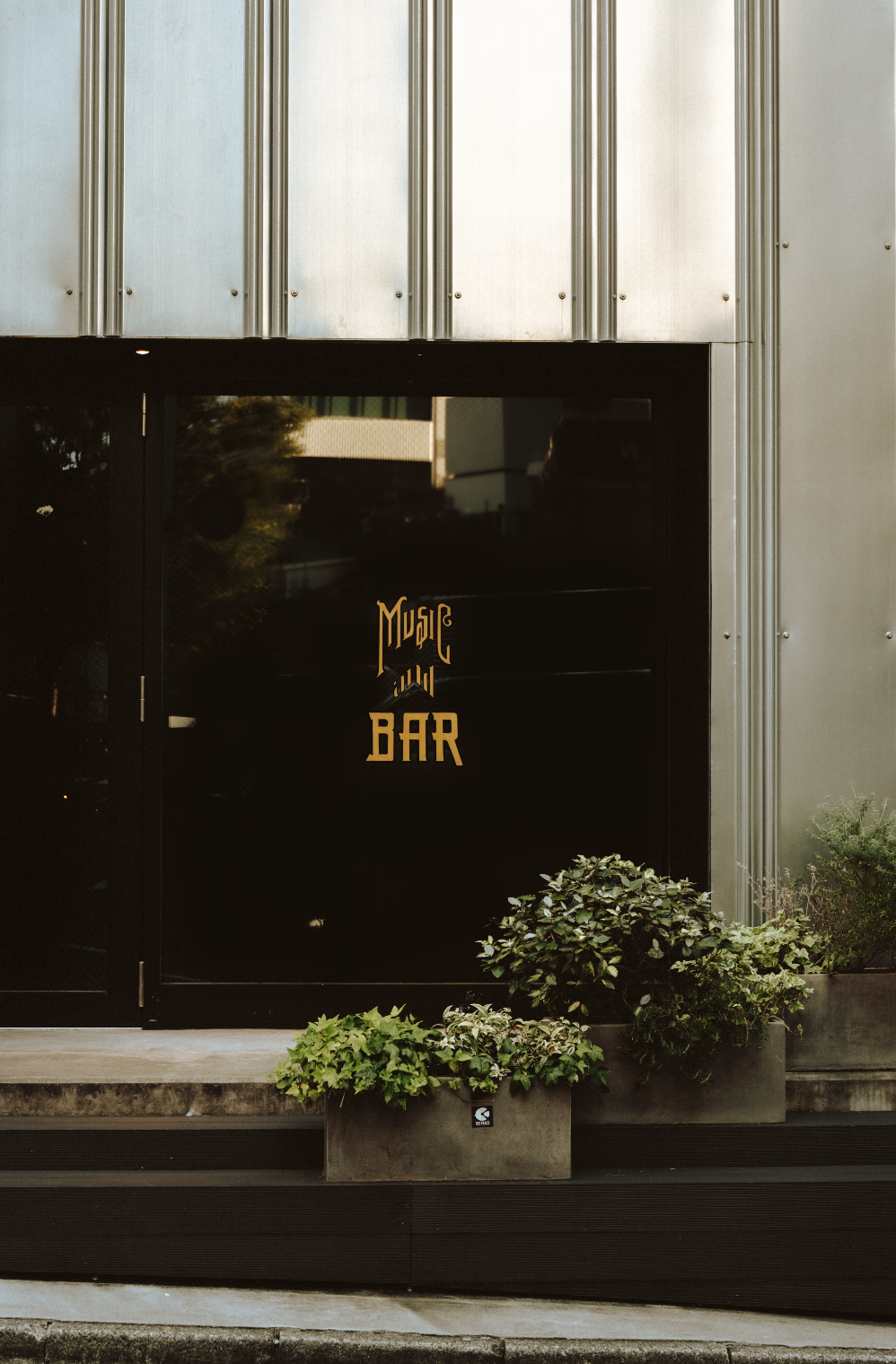 Black exterior door with gold lettering that says 'Music on Bar', flanked by potted plants at the entrance of a building.