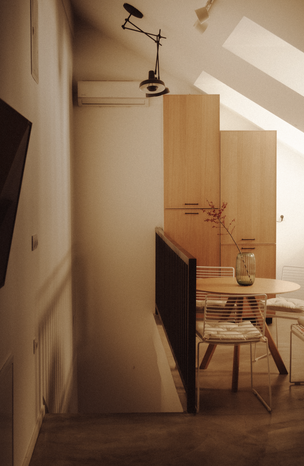 Interior of a modern room featuring a wooden cabinet, a round dining table with a glass vase holding red branches, around four white chairs, a black railing, and a ceiling-mounted TV and light fixture.