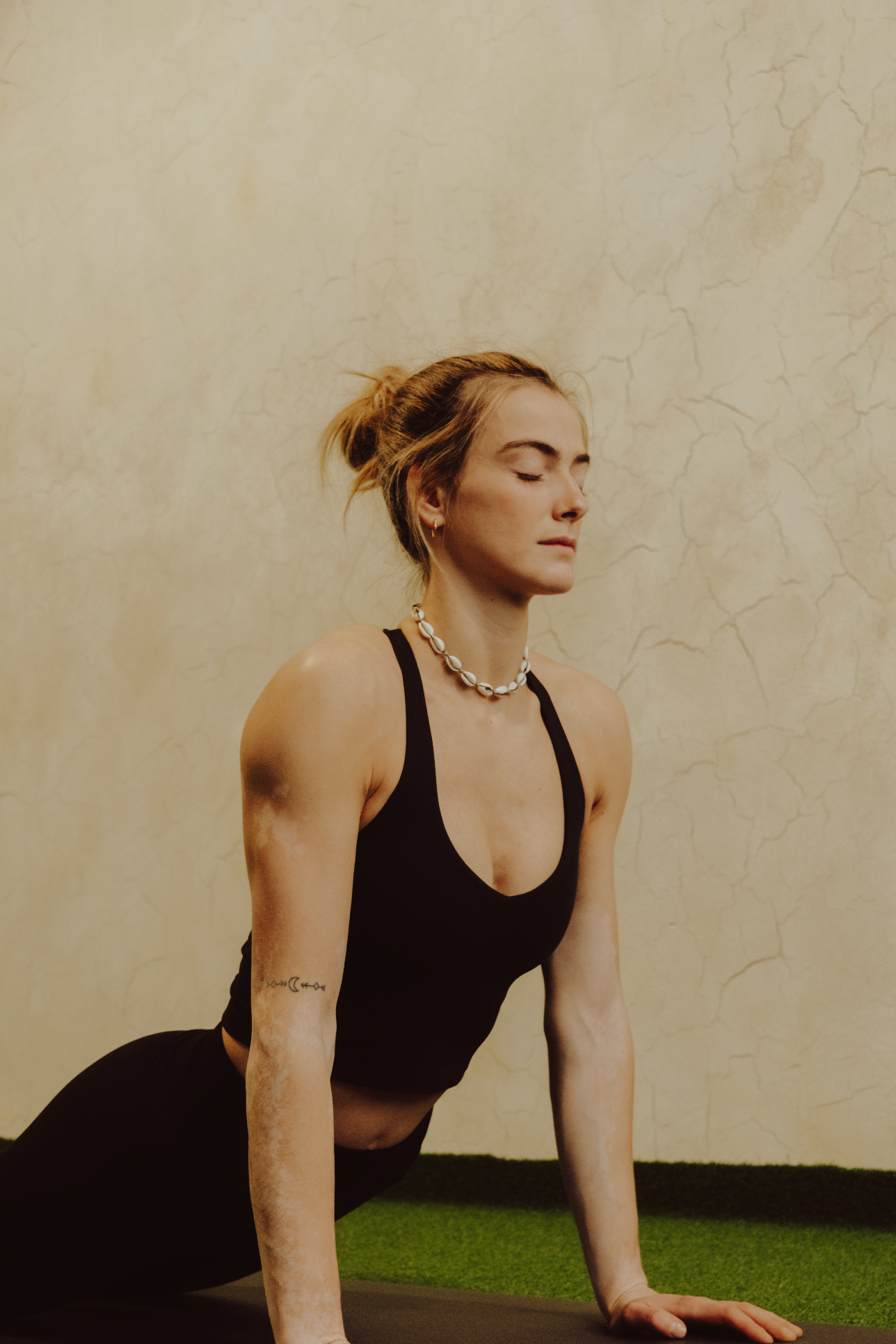 A woman practicing yoga in a plank pose with her eyes closed, wearing a black sports bra and a pearl necklace, on a yoga mat indoors against a beige textured wall.