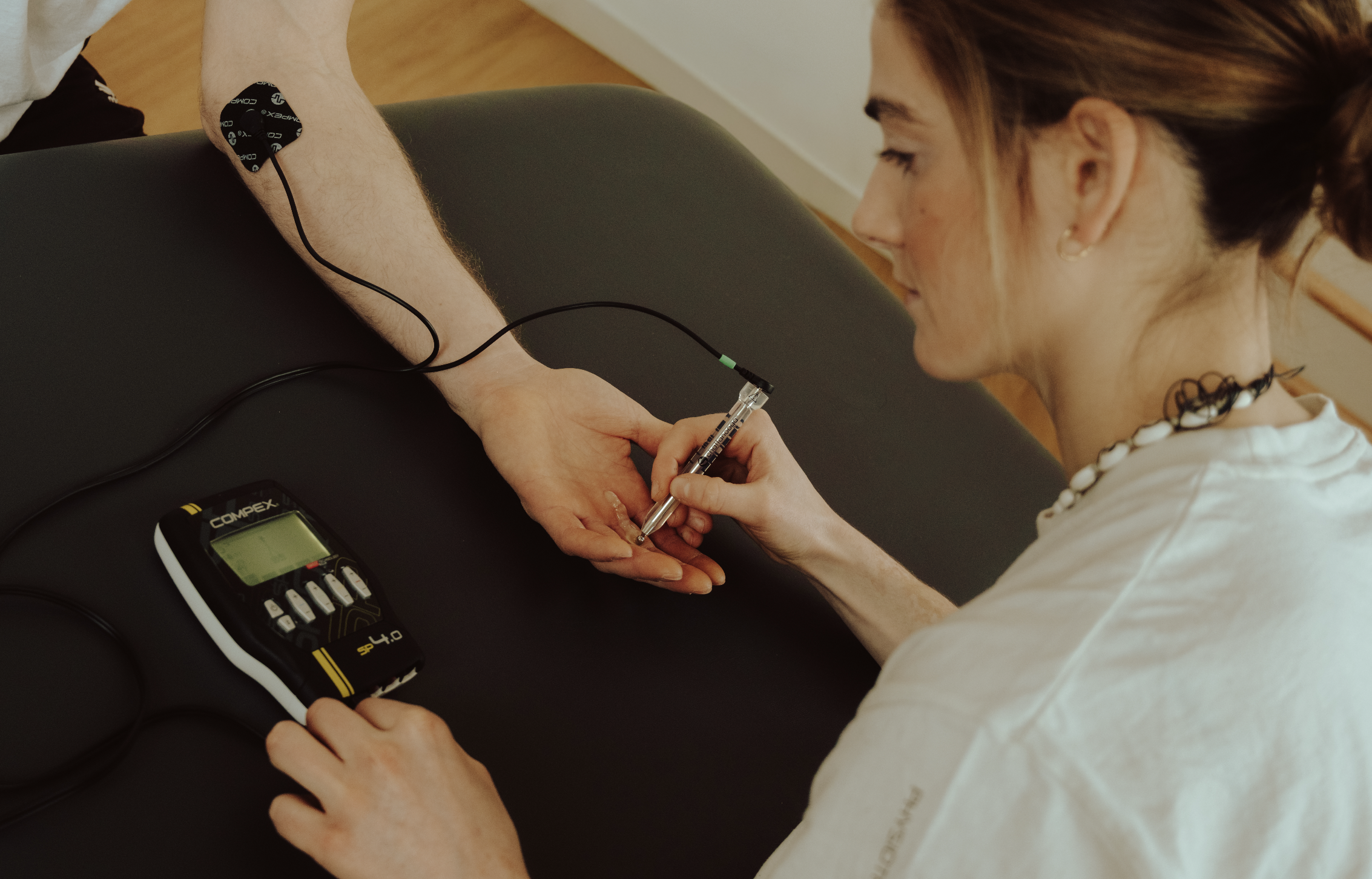 A woman with reddish-brown hair and light skin is receiving a medical injection in her arm from a healthcare worker. The healthcare worker's hand is steady as they insert a needle into her arm. A timer or device is visible on a black surface nearby.
