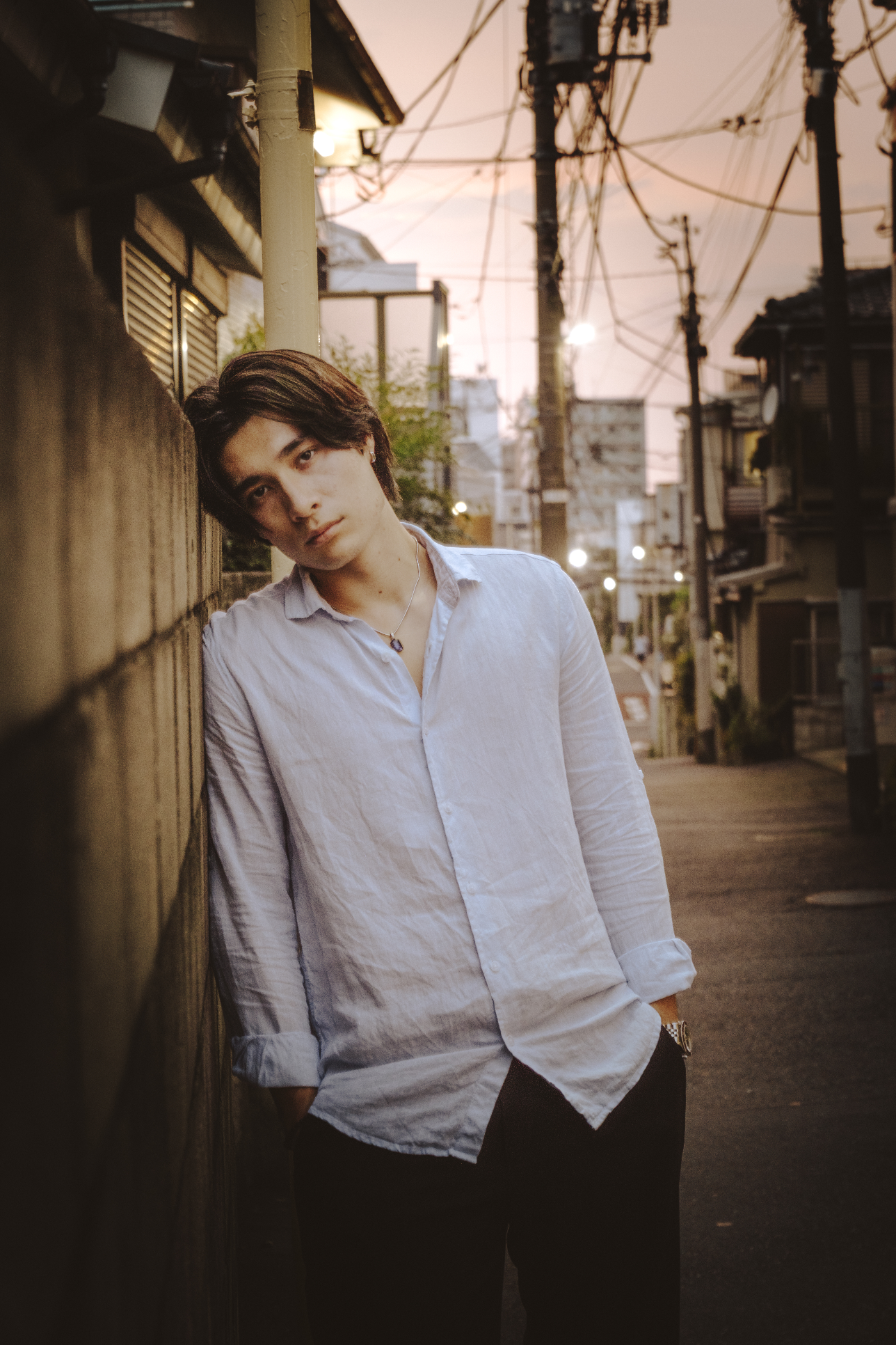 A young man with dark hair leaning against a wooden fence at dusk in an urban alley, wearing a white button-up shirt with rolled-up sleeves and black pants, with a neutral expression.