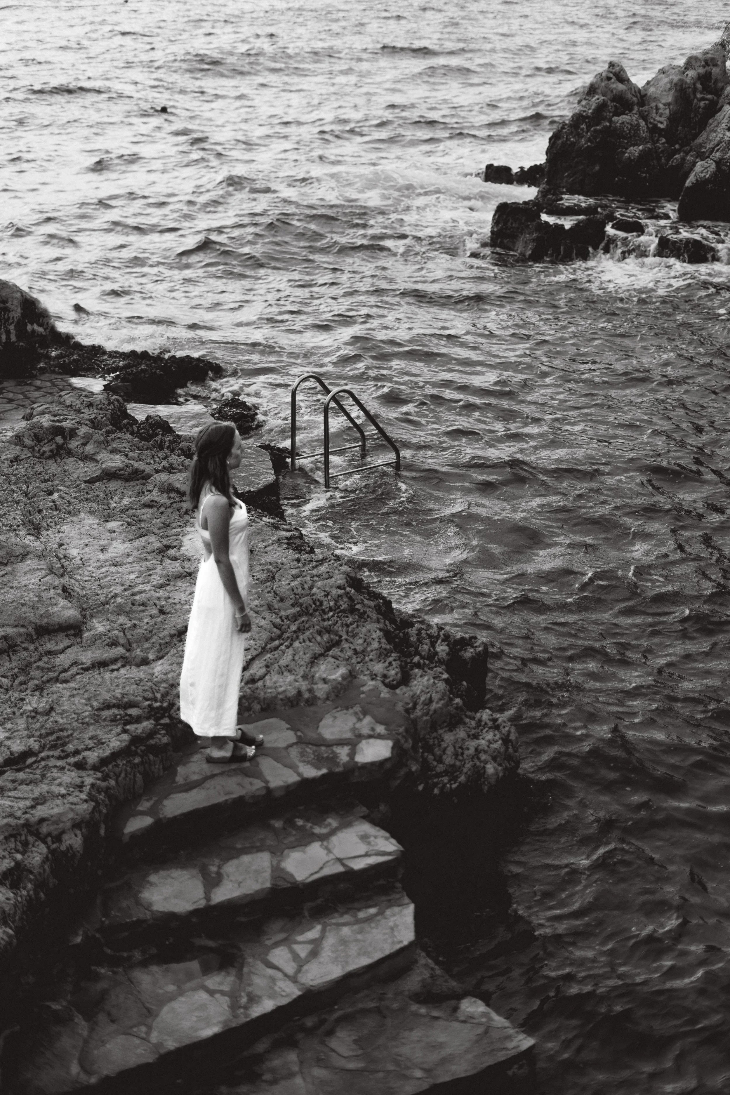 A woman in a white dress standing on rocks near the ocean, with a metal ladder leading into the water, along a rocky shoreline.