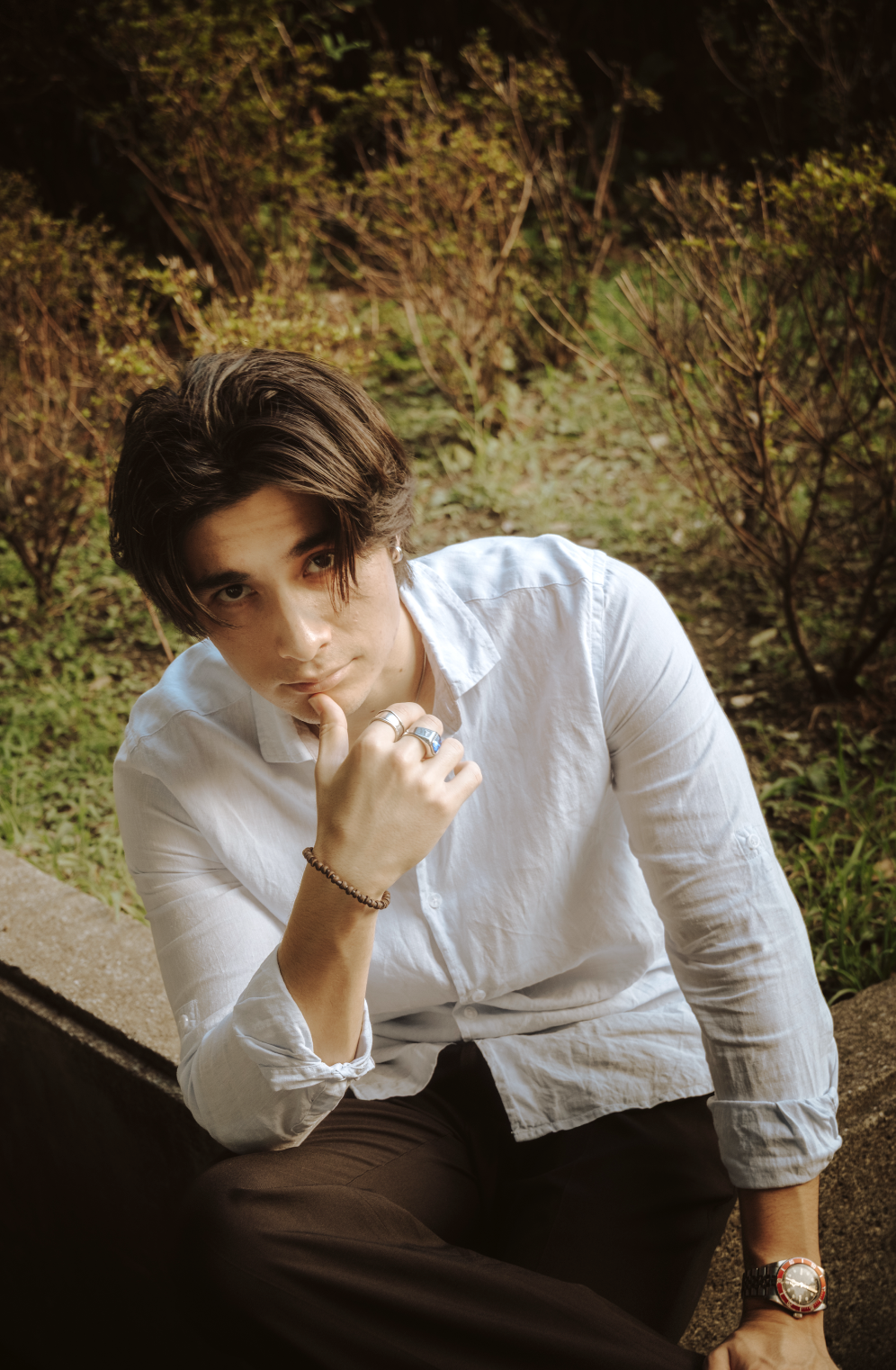 Young man with dark hair, wearing a white button-up shirt and rings, sitting outdoors on a concrete surface, with greenery and trees in the background, looking at the camera with a thoughtful expression.