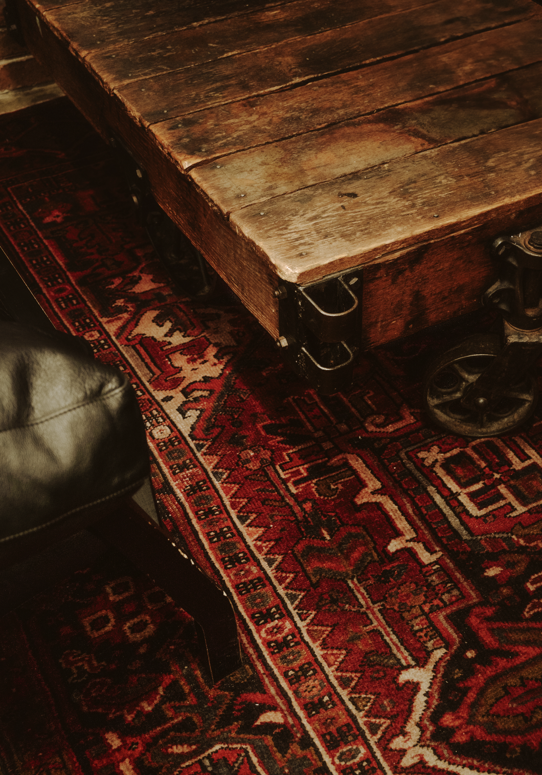 Close-up of a wooden table with caster wheels on a red patterned rug, partially showing a black leather chair.
