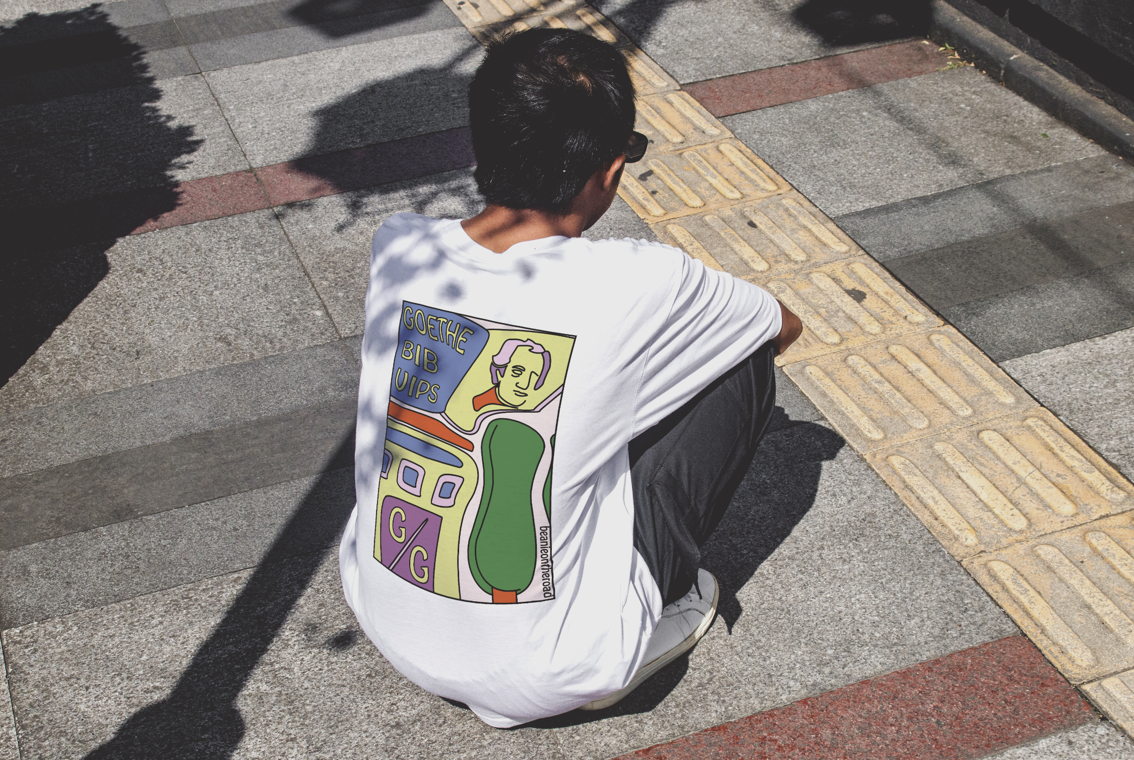 A person with short dark hair wearing sunglasses and a white T-shirt featuring a colorful, artistic design, sitting on a sidewalk with a tiled tactile paving surface, casting a shadow in the sunlight.