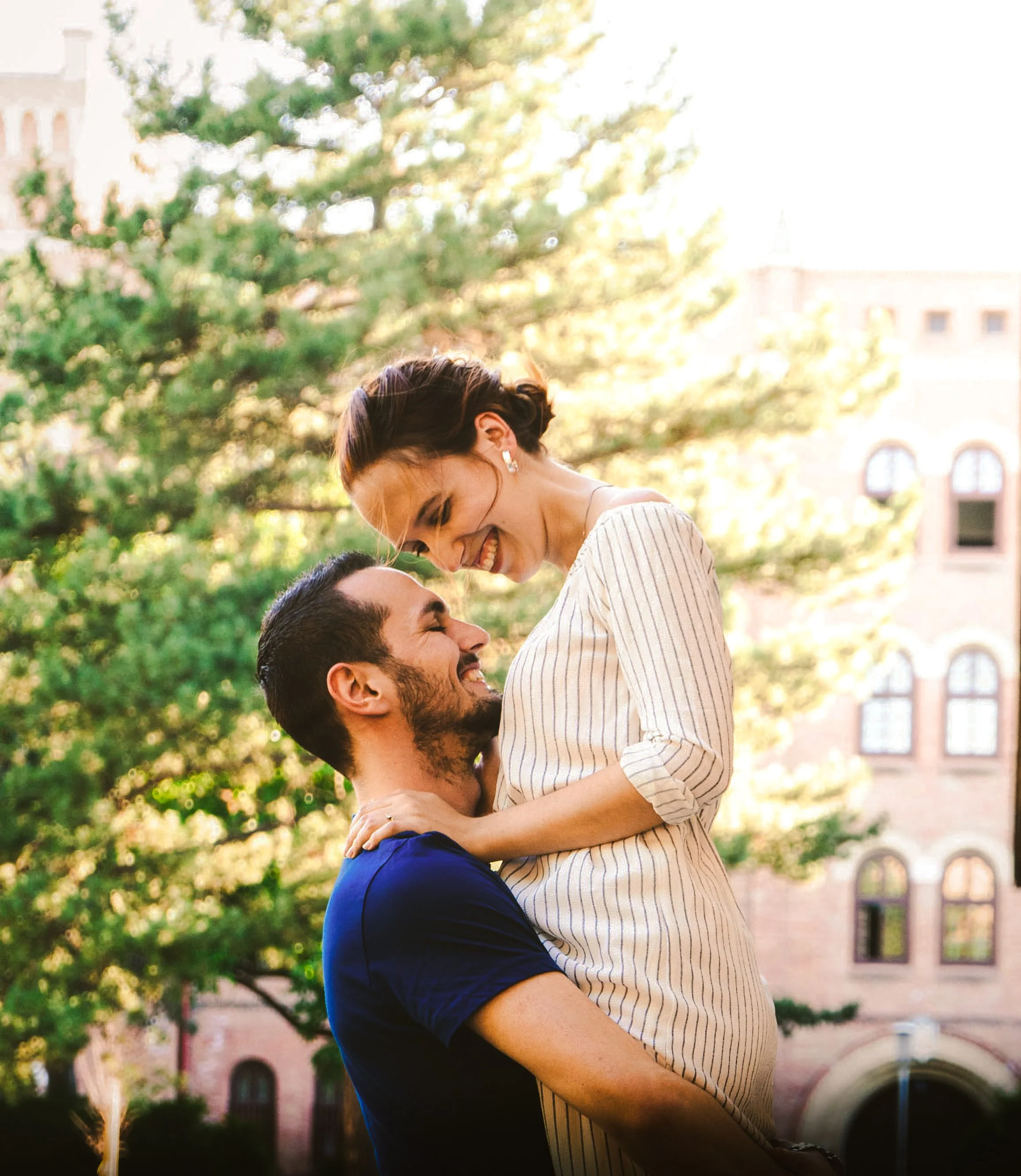 A smiling couple with the man lifting the woman in an outdoor park setting, with trees and buildings in the background during daytime.